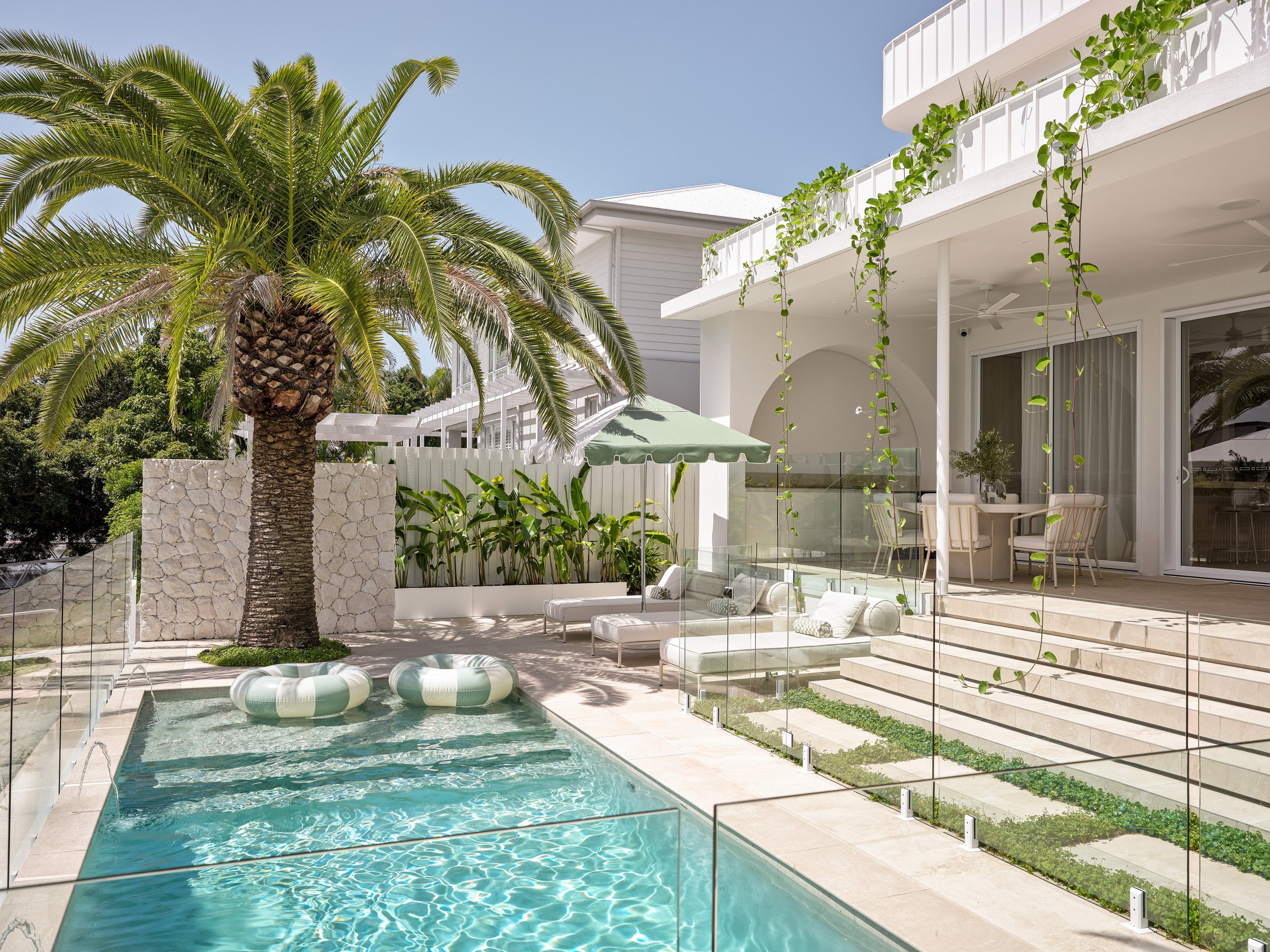 Modern white house with a backyard pool, palm tree, outdoor furniture, and green plants under a clear blue sky.