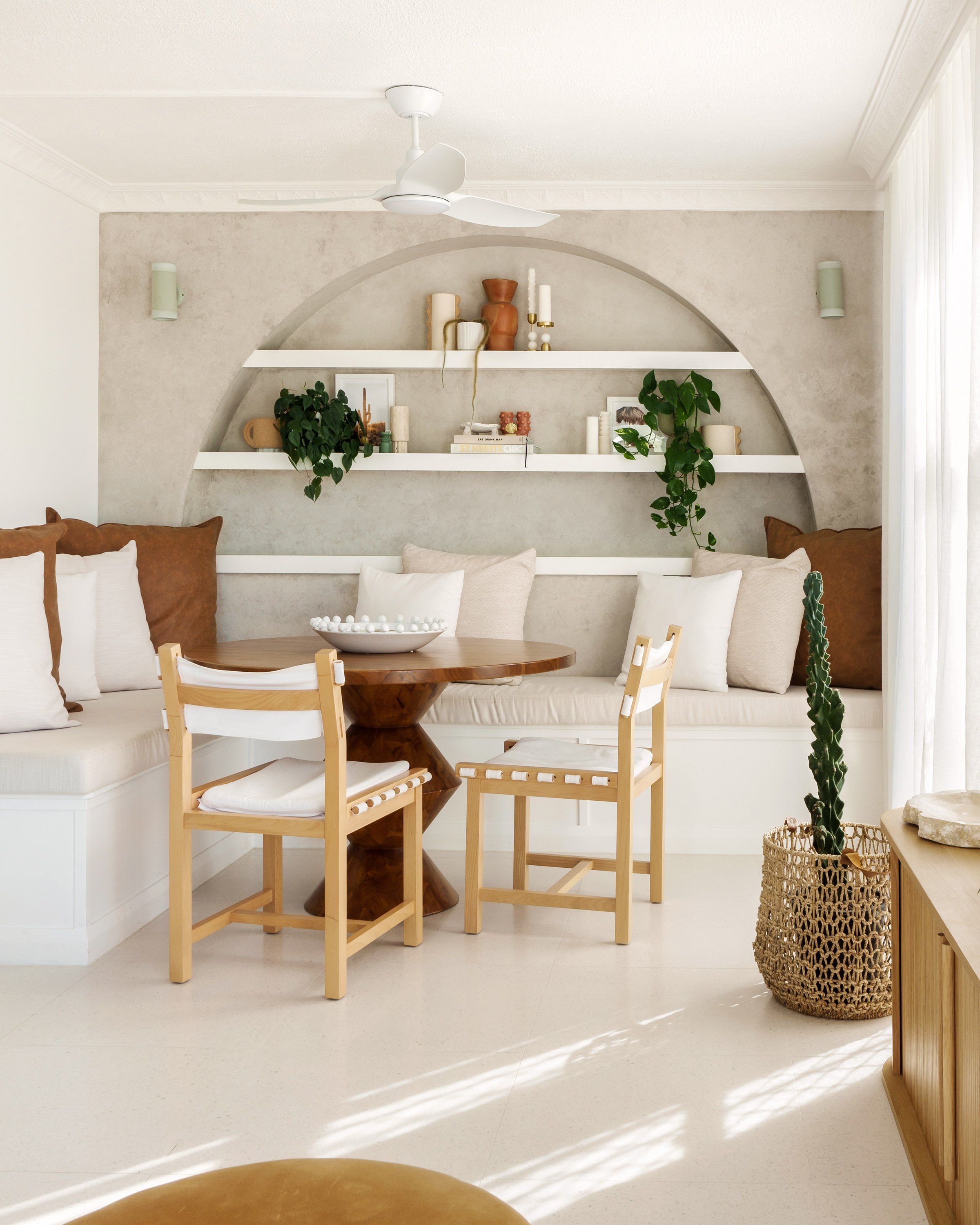 A cozy dining nook with beige and brown cushions on built-in benches, a round wooden table with a decorative bowl, and two light wood chairs with white cushions. The background features a geometric wall with two shelves holding books, candles, and de