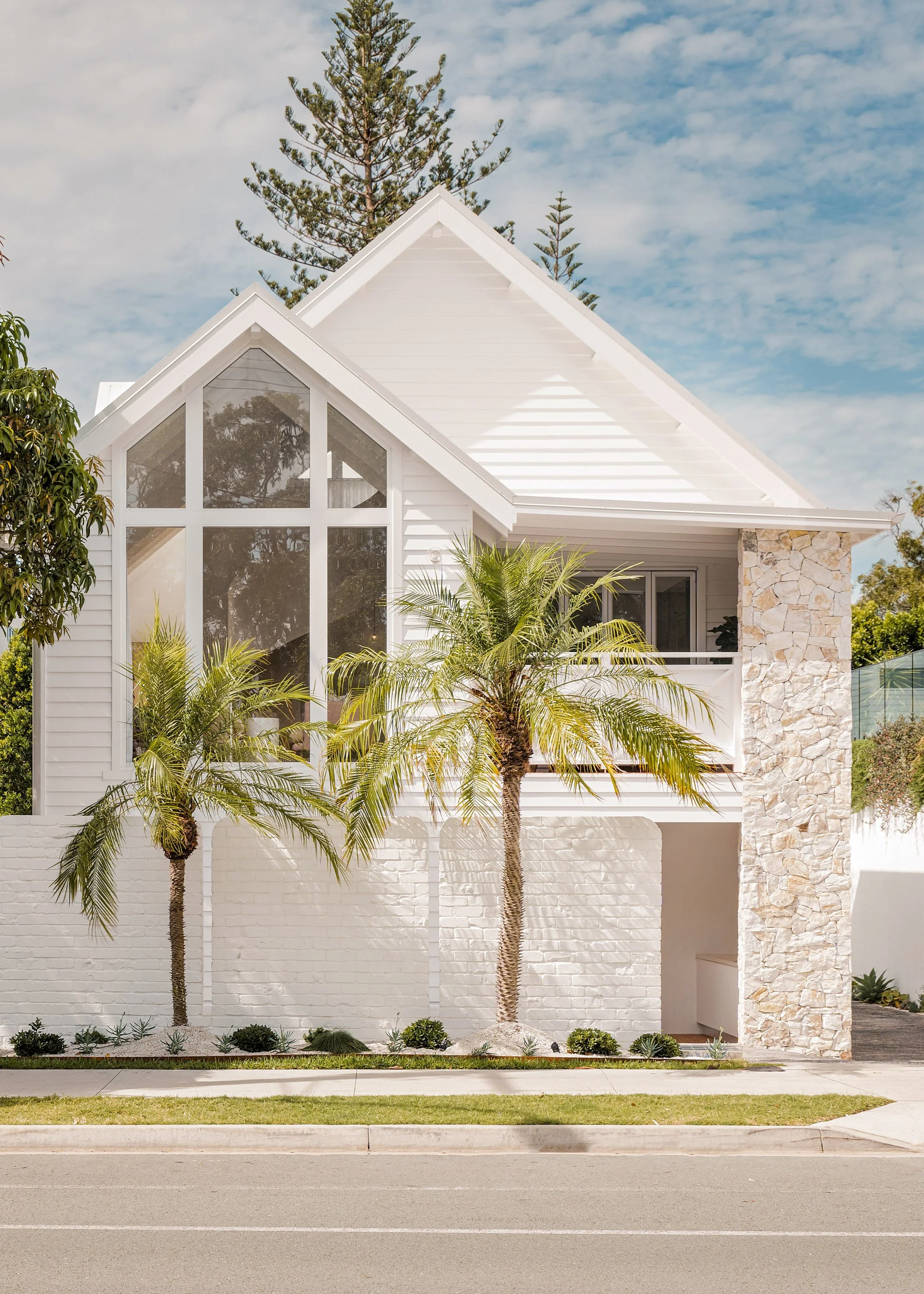 Modern white house with large glass windows, stone accent wall, and palm trees in front, under a partly cloudy sky.