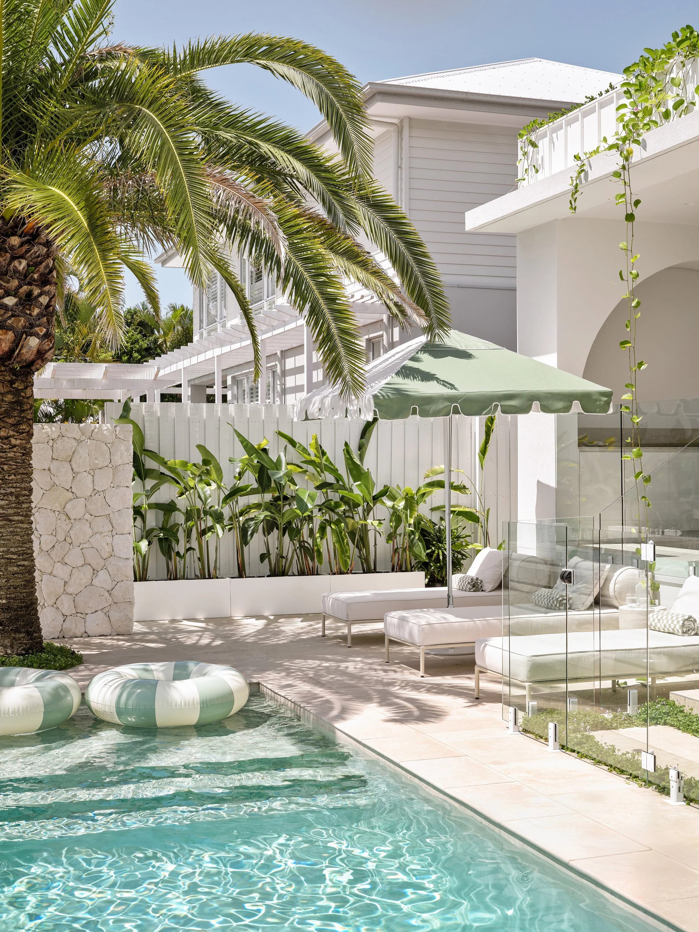 Poolside lounge chairs with cushions, green palm tree, large house in background, pool floats in water, white patio furniture, green patio umbrella, glass pool fence, lush green plants, sunny day