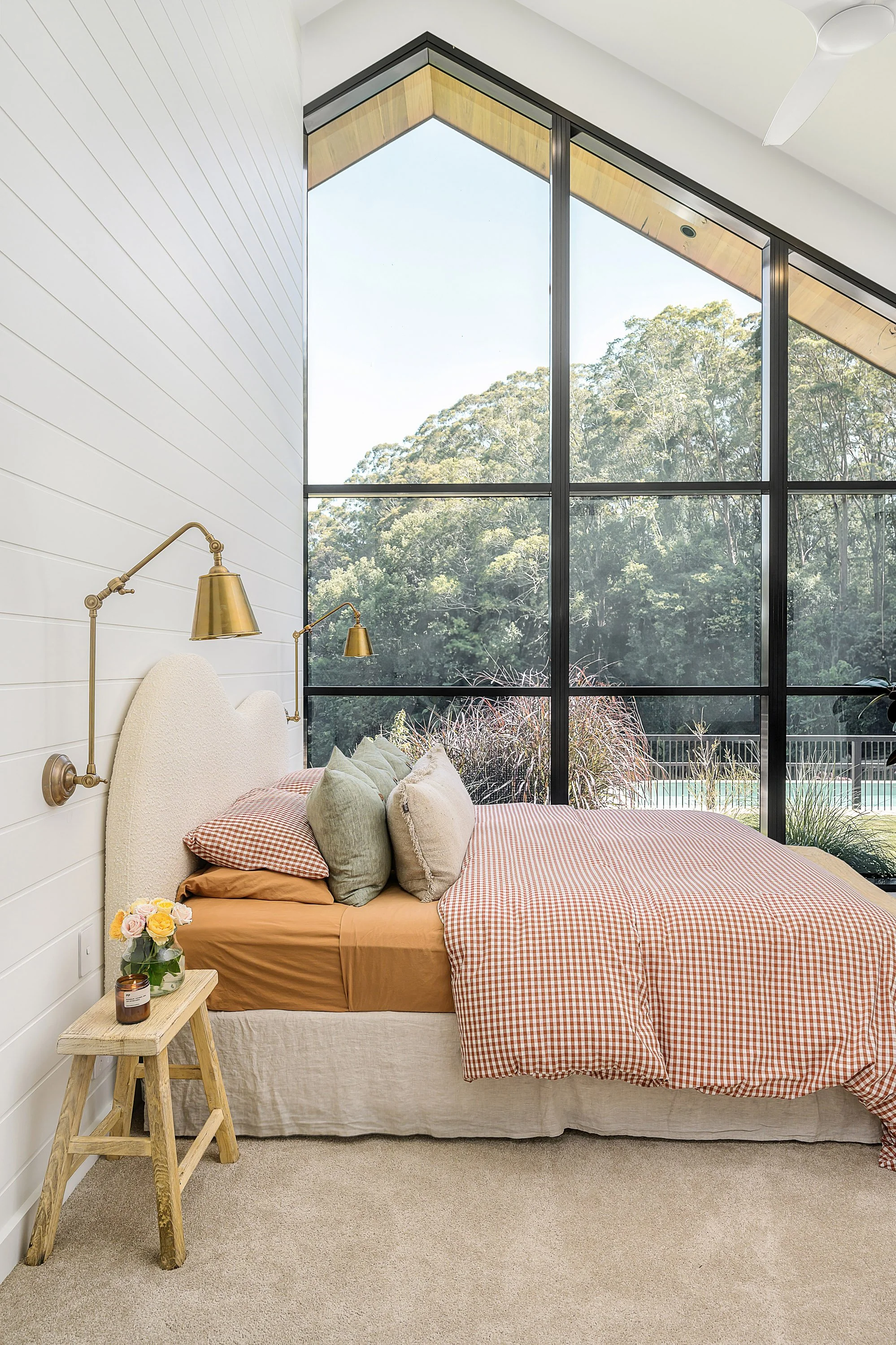 Bedroom with large floor-to-ceiling windows showing trees outside, with a bed covered in orange and checkered bedding, beige and green pillows, a small wooden nightstand with a flower vase, and gold wall-mounted reading lamps.