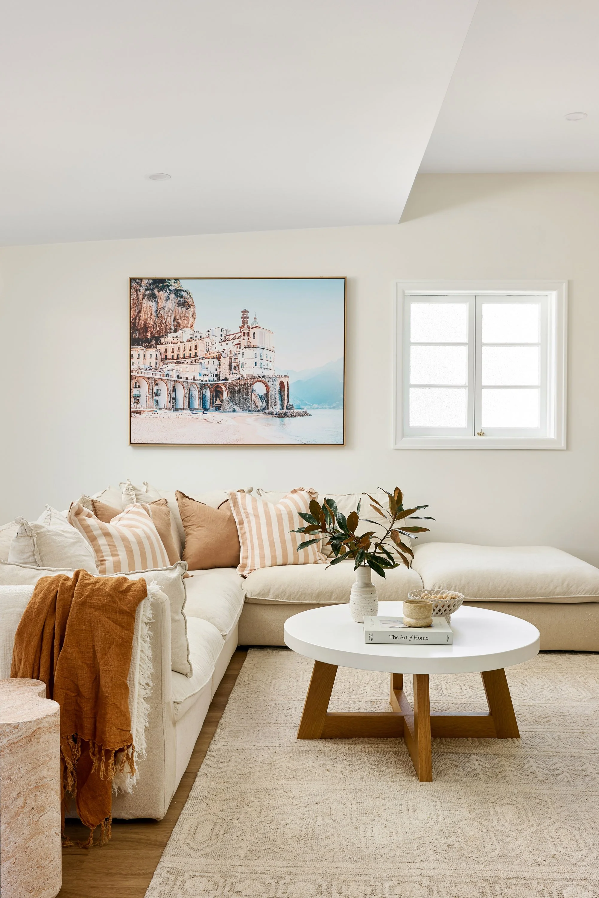 Bright living room with beige sectional sofa, striped and solid throw pillows, a round white coffee table with a potted plant, decorative bowl, and book, cream-colored rug, window, and balcony with cityscape painting.
