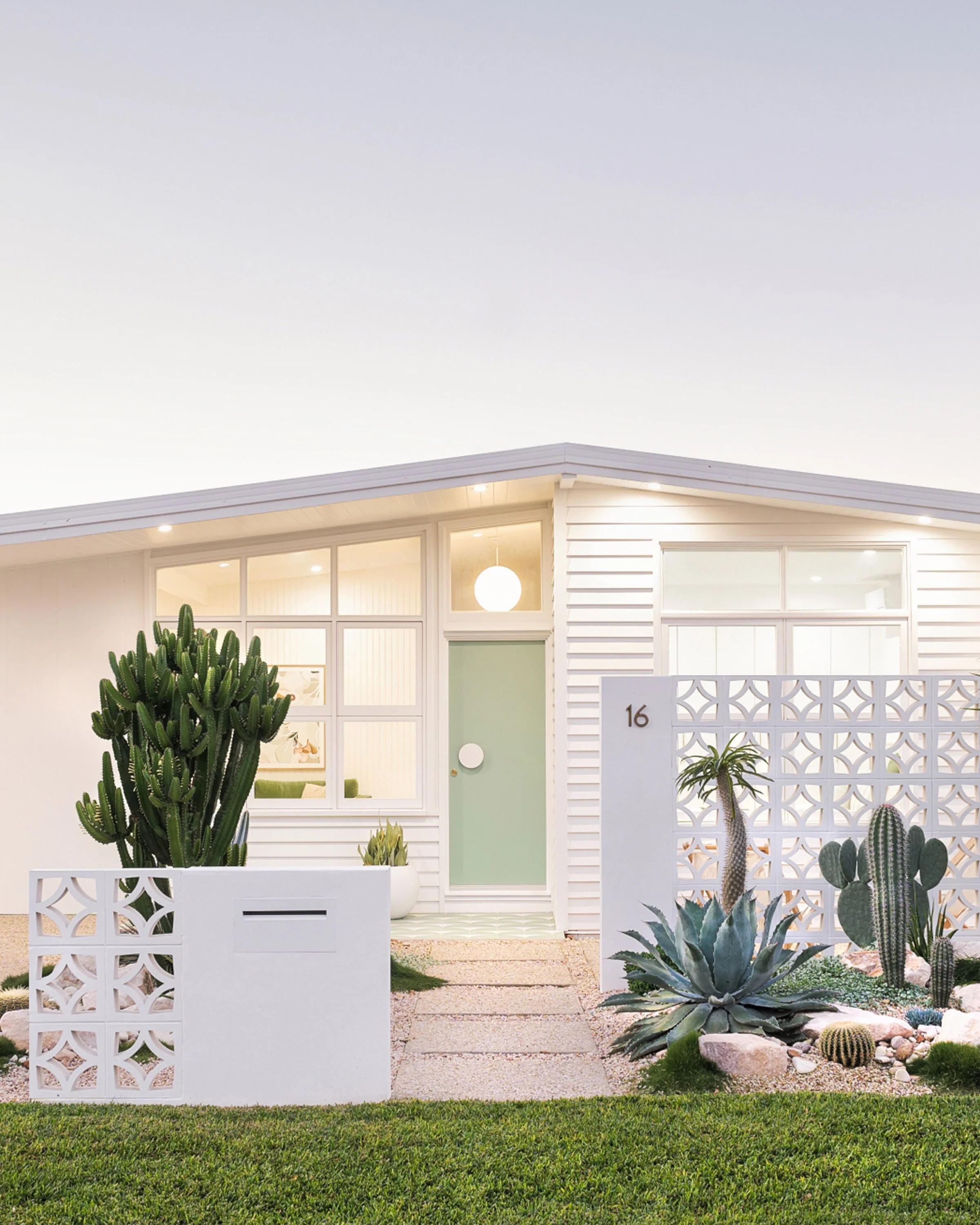 A modern white house with large windows, a mint green front door, and desert plants including cacti and succulents in the yard, with a white lattice fence and well-manicured lawn.