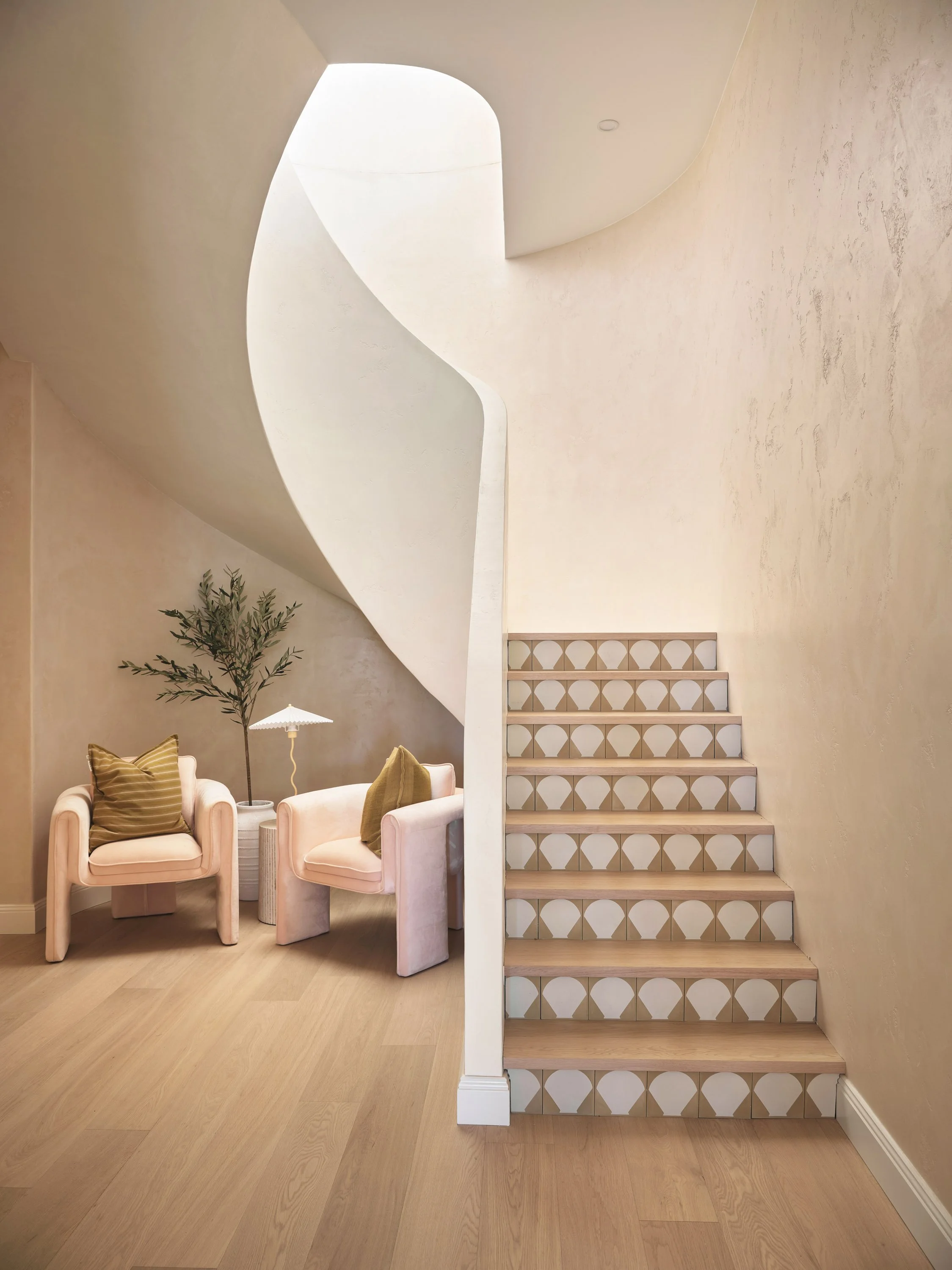 Interior of a modern home featuring a curved staircase with geometric patterned risers, two pink armchairs with striped and solid cushions, a small table lamp, a potted plant, and light wooden flooring.