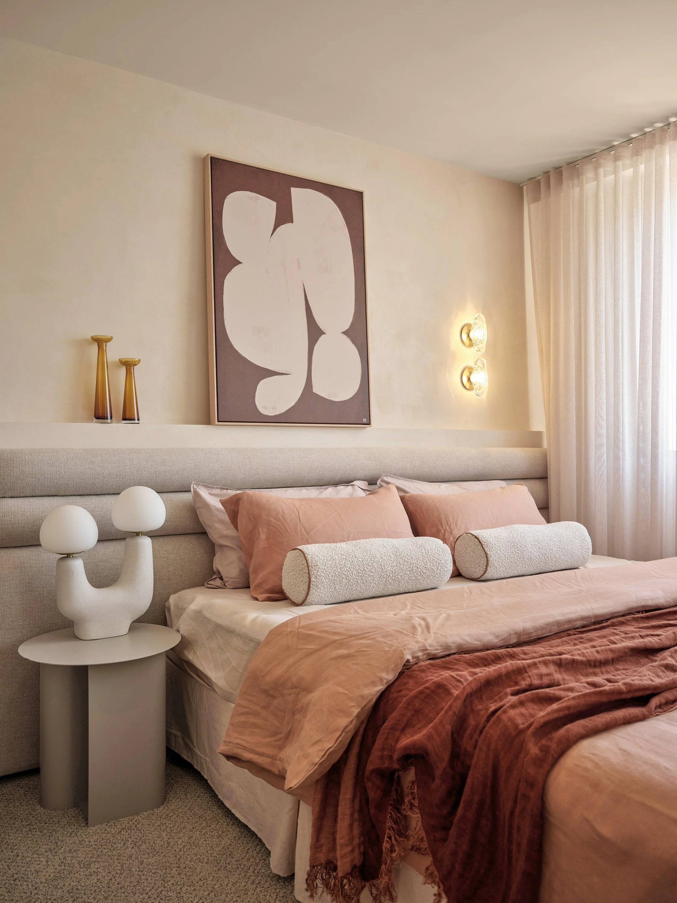 A neatly made bedroom with a bed decorated with pink and beige bedding and matching pillows, a beige upholstered headboard, a modern lamp on a small side table, pendant lights on the wall, abstract artwork above the bed, and curtains covering a large