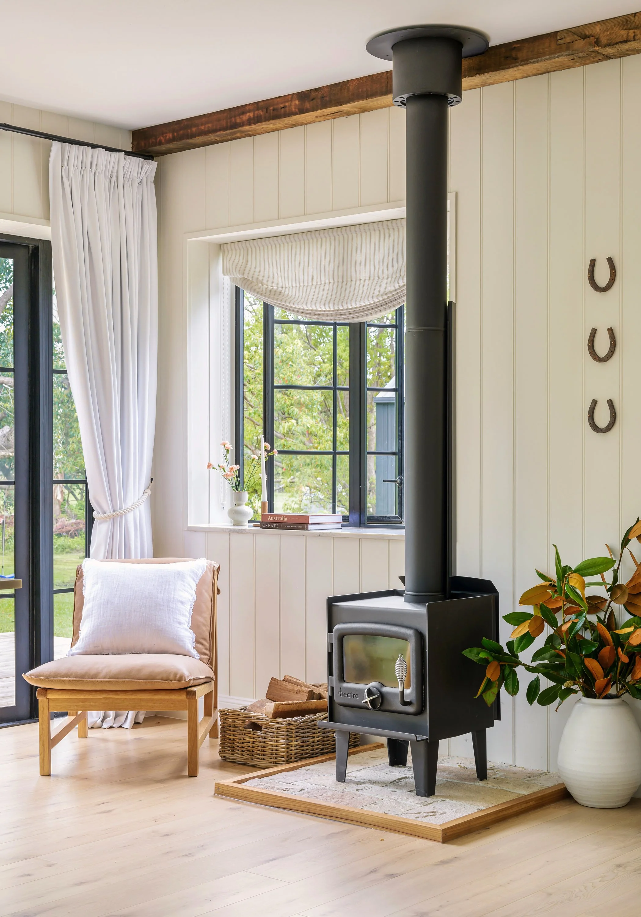 Living room corner with a wood stove, a beige chair with white pillow, a basket of firewood, a large potted plant, a window with white curtains, and black-framed glass door, with decorative horseshoes on the wall.