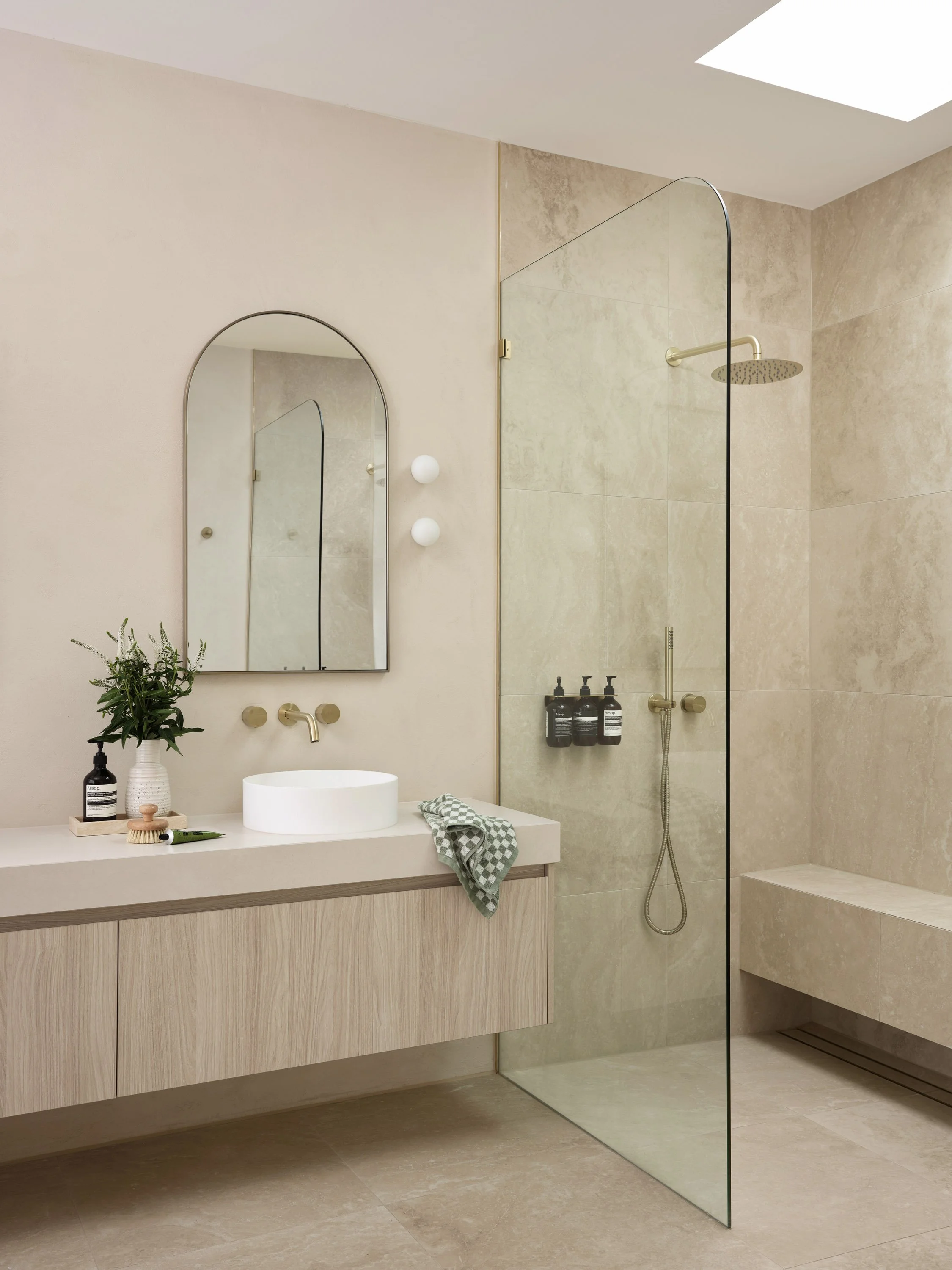 Modern bathroom with beige tiles, a wooden vanity with a round white basin sink, a mirror, and a glass-enclosed shower area with a rain shower head and black bottles on a wall-mounted dispenser.