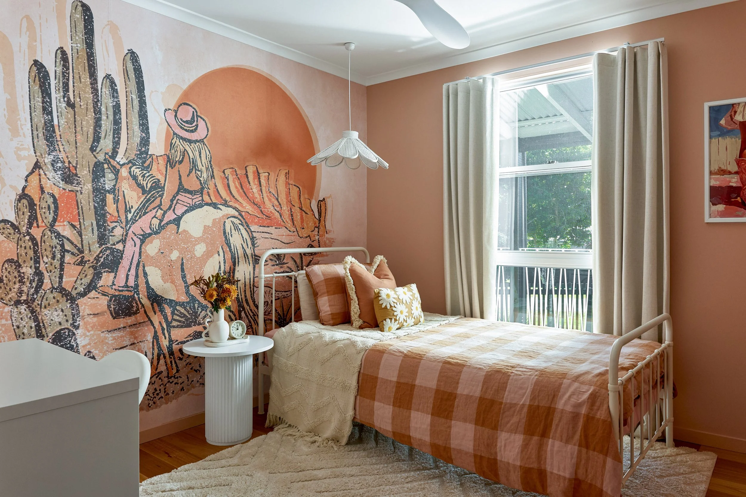 Bedroom with pink walls, a mural of a woman riding a horse among cacti on one wall, white bed with checkered pink and orange bedding, pillows, a white side table with a vase of flowers, a window with white curtains, and a ceiling fan.