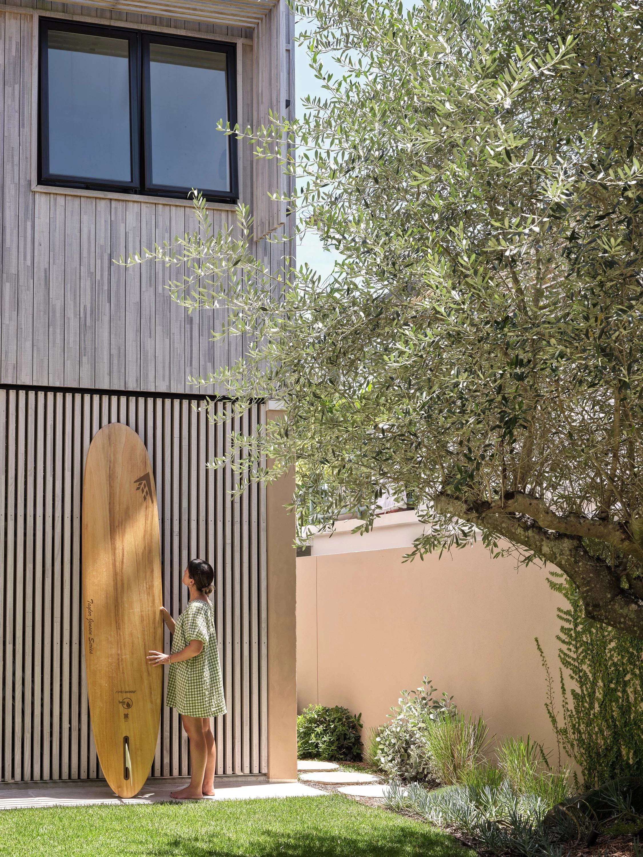 A woman in a green checkered dress standing outside next to a wooden surfboard, touching it, with a modern house and lush garden in the background.