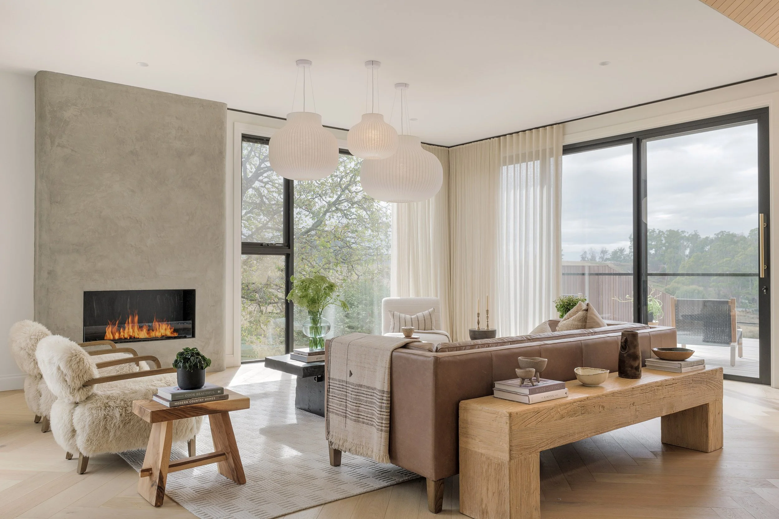 Modern living room with a fireplace, large windows, white curtains, and a beige sectional sofa with decorative pillows. There are two plush armchairs, a small wooden side table with books and a plant, and a wooden bench with decorative objects. Three