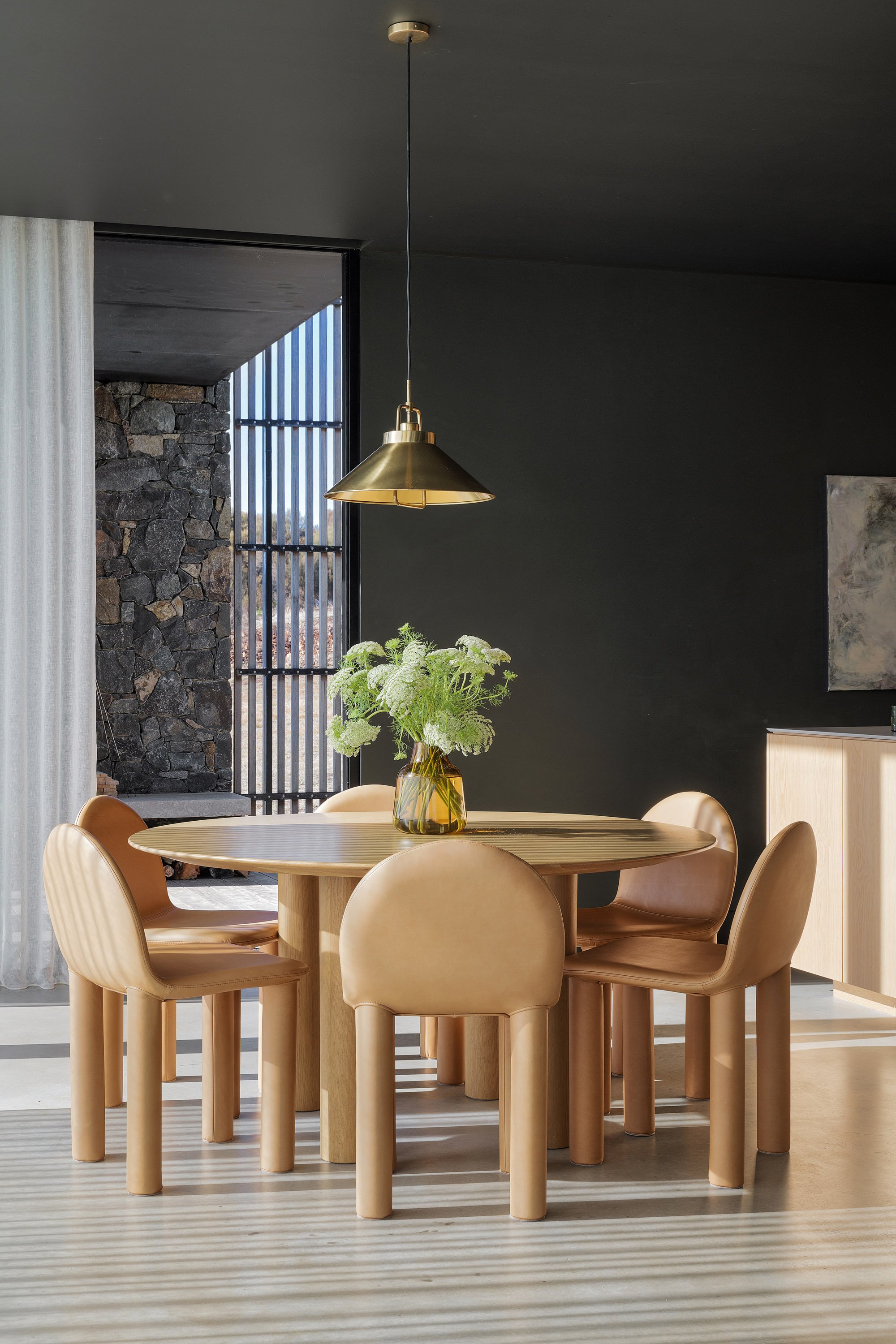 A dining room with a round wooden table, six beige chairs, a vase with green flowers, a brass pendant light, dark wall, stone wall outside the window, and natural light streaming in.