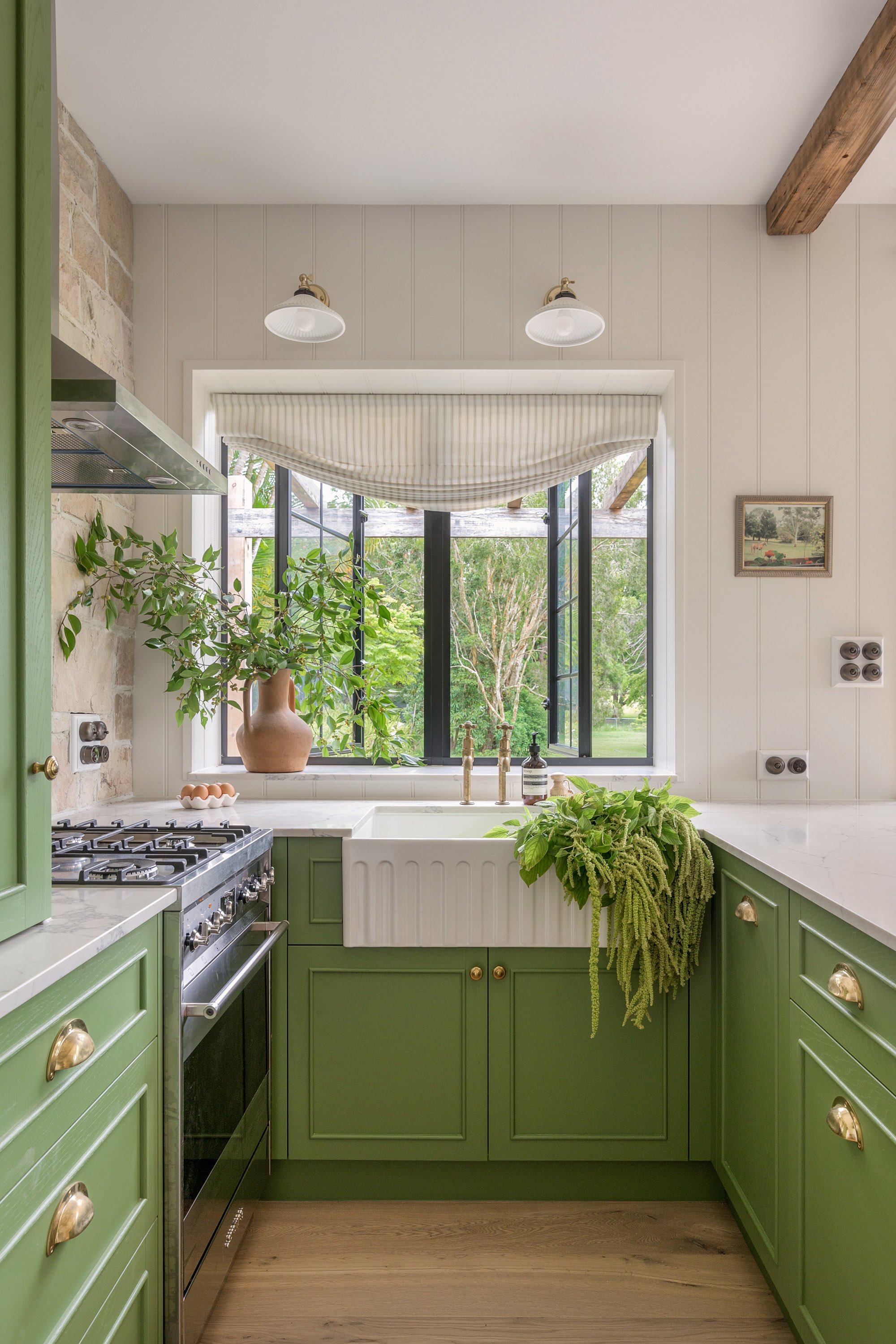 Kitchen with green cabinets, white marble countertops, window with striped valance, and garden view outside.