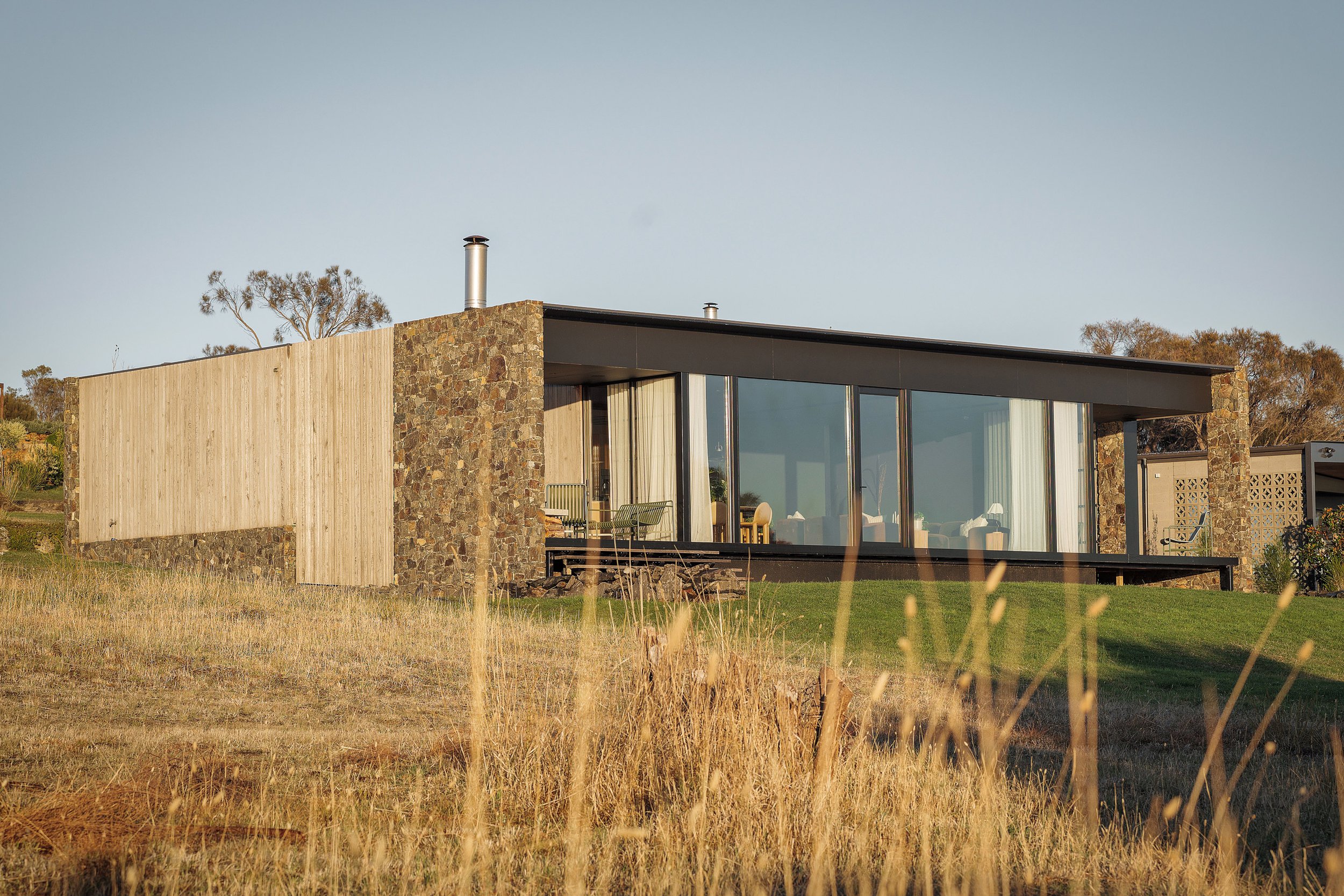 Modern house with large glass windows, stone and wood exterior, situated on a grassy hillside with a clear sky and trees in the background.