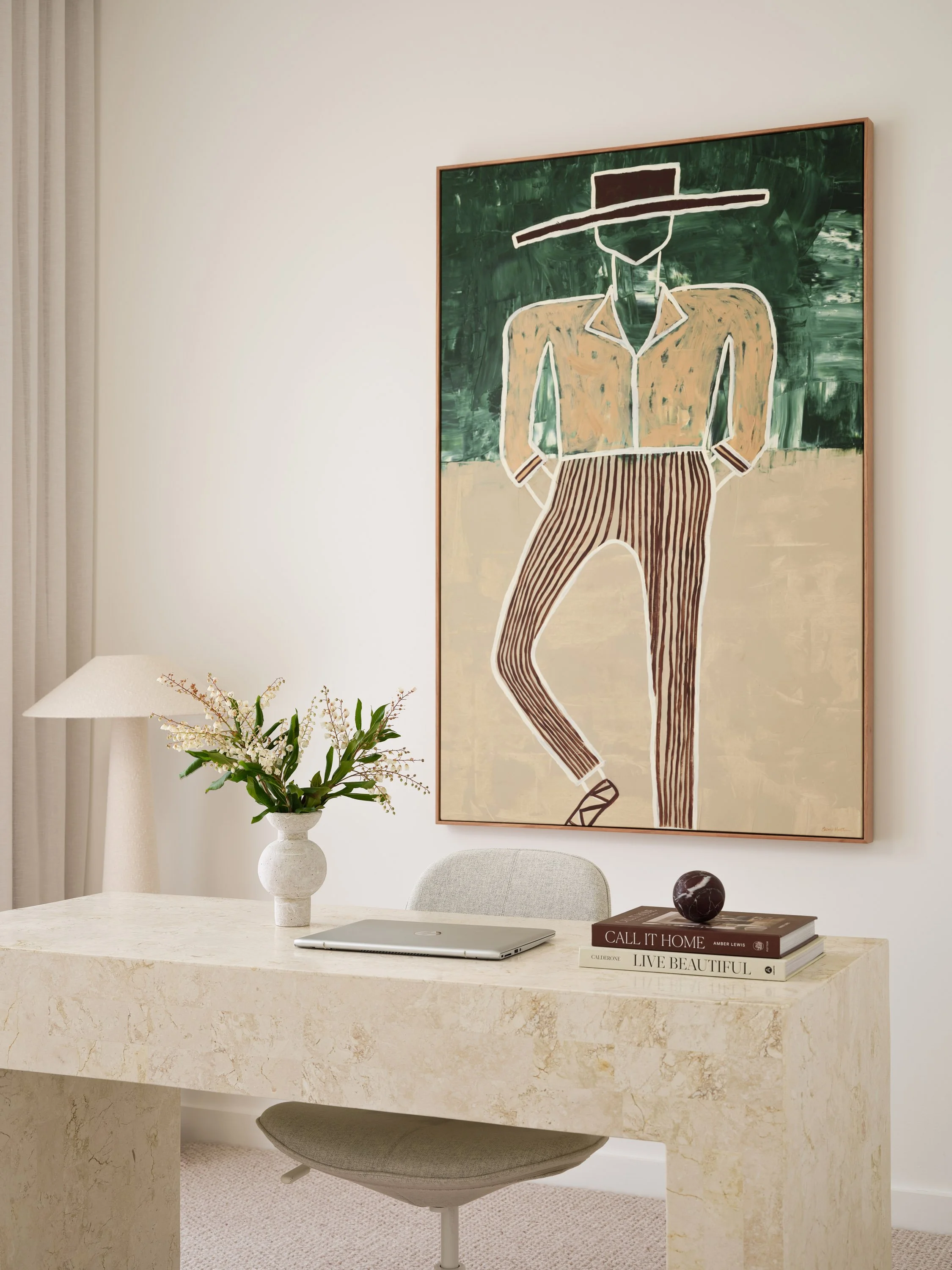 Modern minimalist home office with a marble desk, a gray office chair, a table lamp, a vase with white and green flowers, and a large abstract painting of a person with a wide-brimmed hat hanging on the wall.