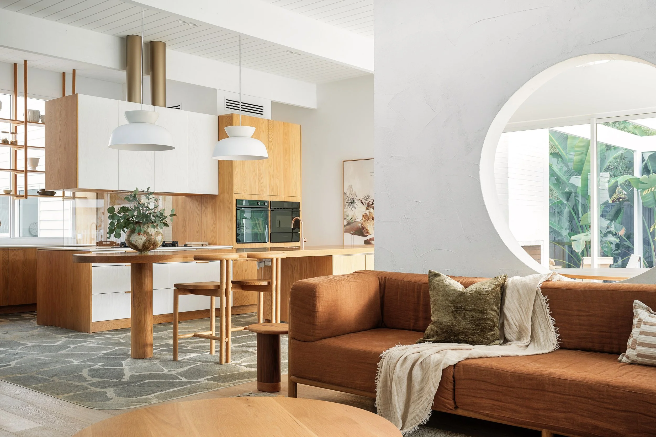 Modern open-concept living room and kitchen with wooden cabinetry, a brown sofa with cushions, and large windows with greenery outside.