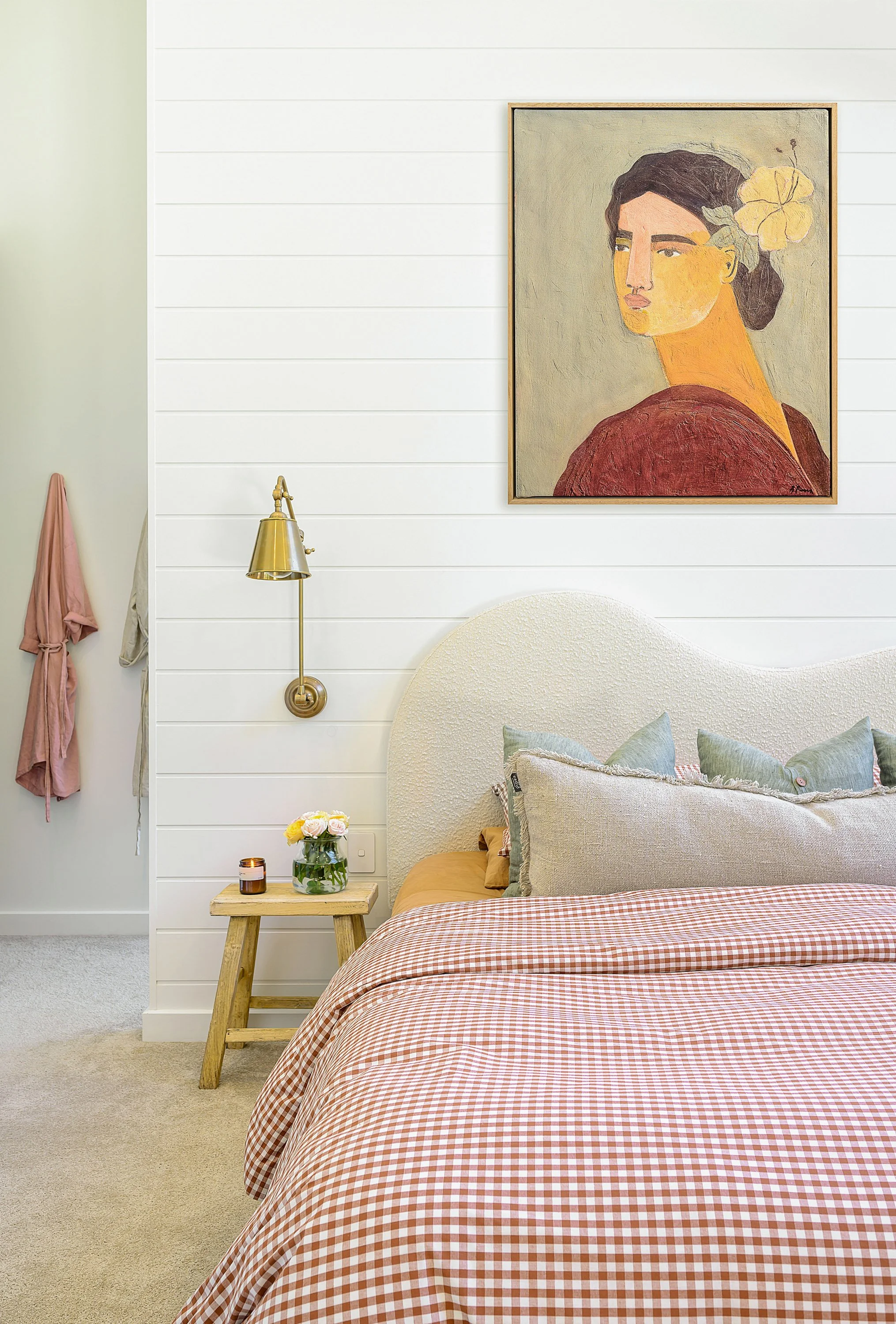 A cozy bedroom with a white textured headboard, a gingham bedspread, several pillows, a small wooden side table with a flower vase and candle, a gold reading lamp, and a framed portrait of a woman with dark hair and a yellow flower in her hair hangin