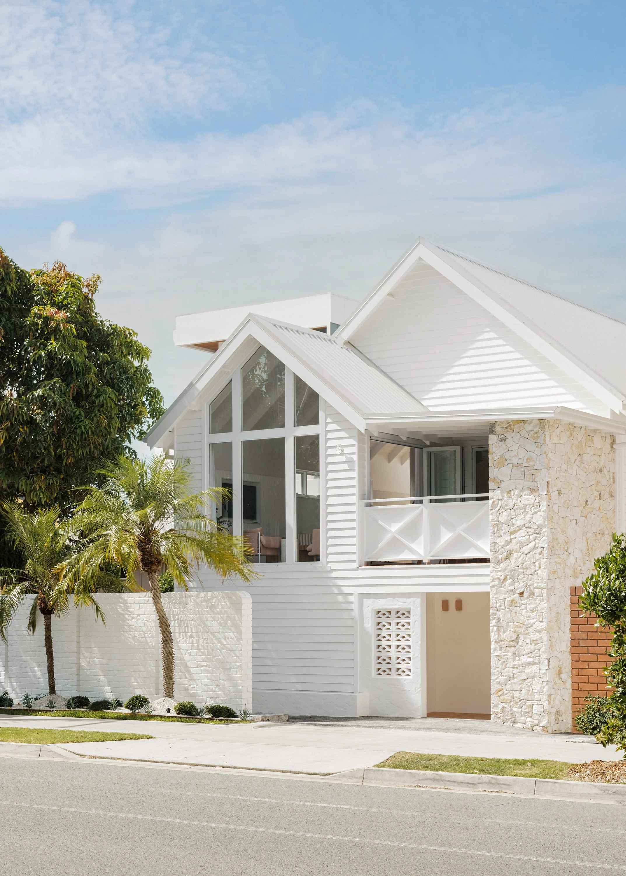 Modern white house with large windows and stone accent wall, surrounded by palm trees, on a street under a blue sky.
