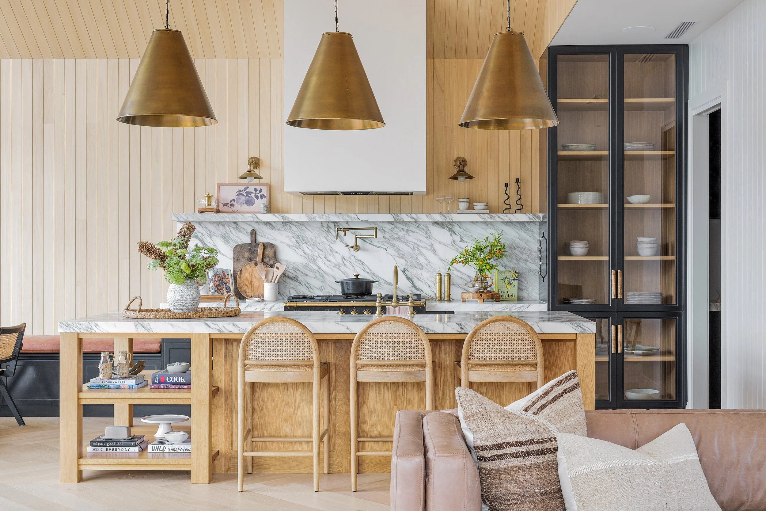 Modern kitchen with wooden cabinets, marble countertops, and black glass-fronted pantry. Three gold pendant lights hang above a breakfast bar with wooden chairs, and there is a brown sofa with throw pillows in the foreground.