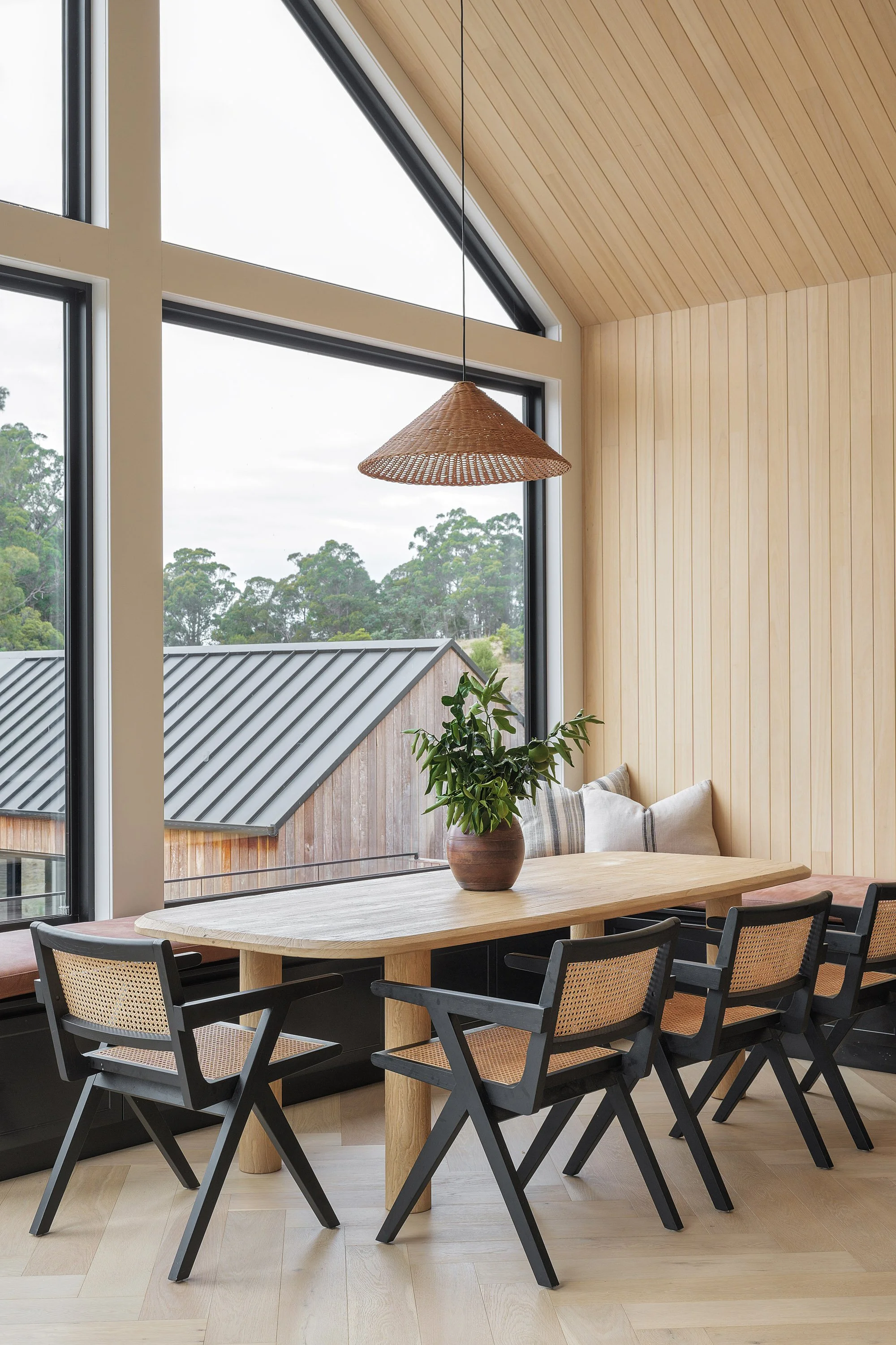Interior of a dining area with a wooden table, black chairs, a window seat with cushions, a potted plant, and wood-paneled walls and ceiling.