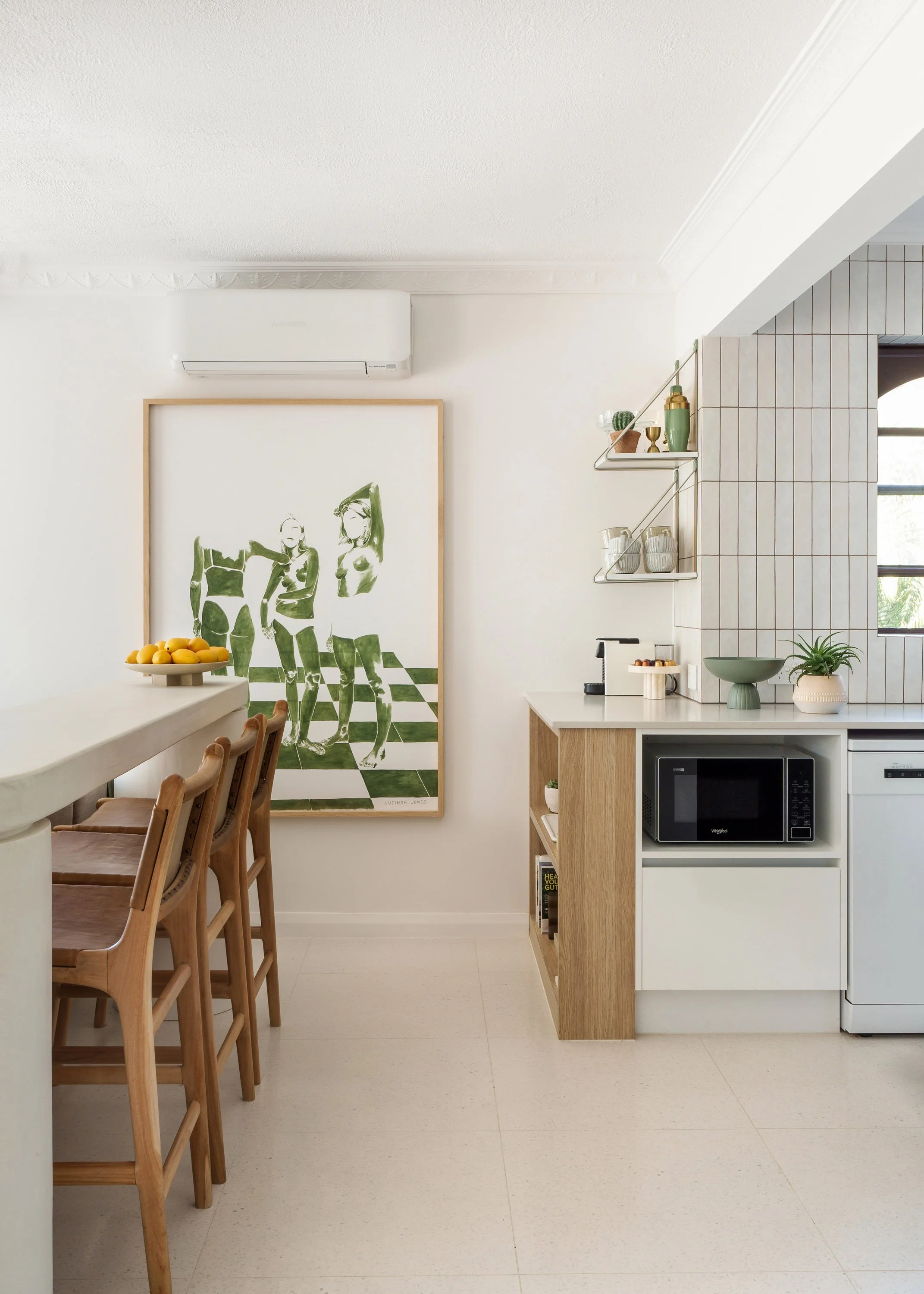 Modern kitchen with wooden chairs, a large framed artwork of three women, a white countertop with a bowl of lemons, shelves with plant and decorative items, and kitchen appliances including a microwave and coffee maker.