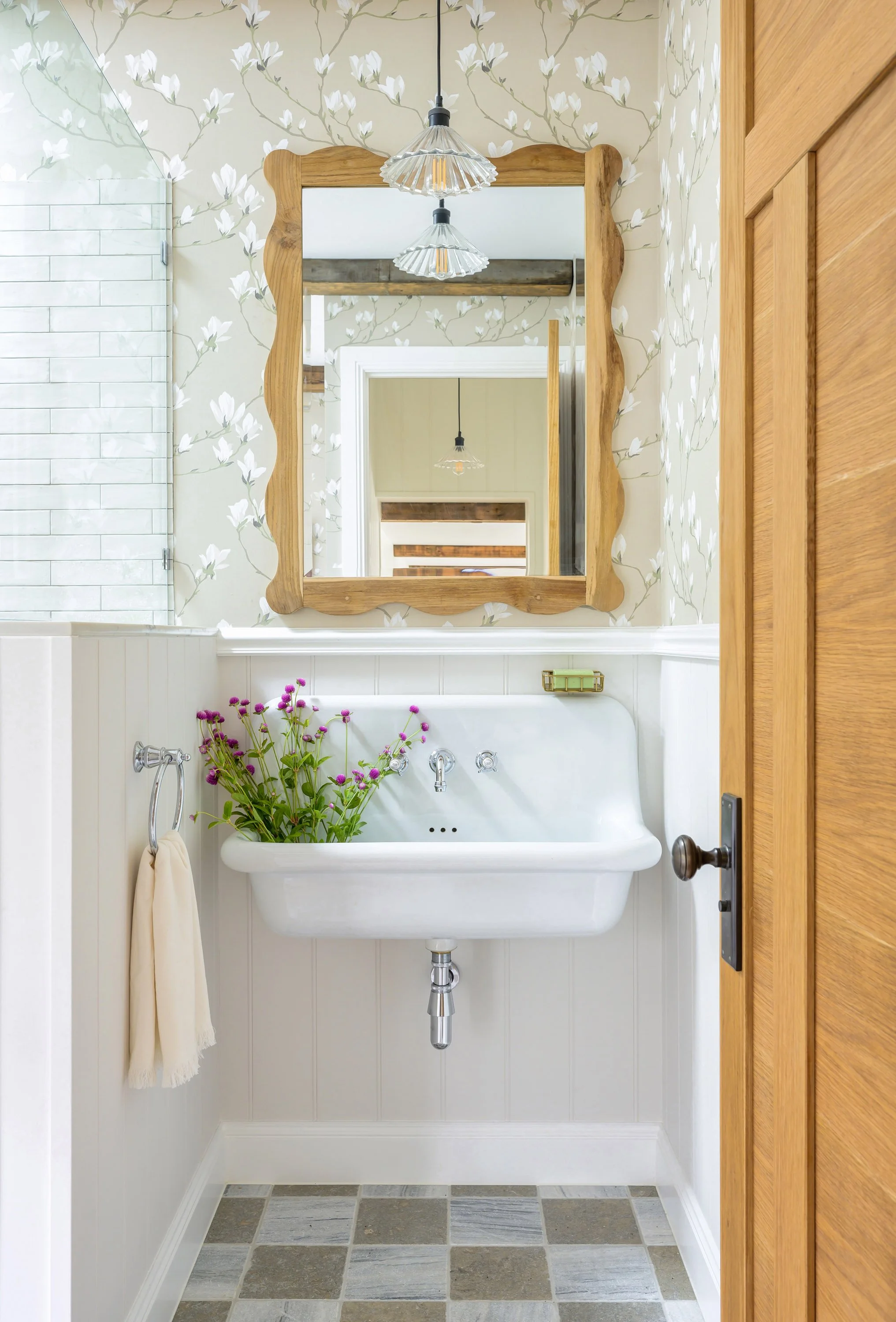 A vintage white sink with chrome fixtures and a potted plant with purple flowers, mounted on a white paneled wall below a wooden-framed mirror, in a bathroom with floral wallpaper, a wooden door, and a tiled floor.
