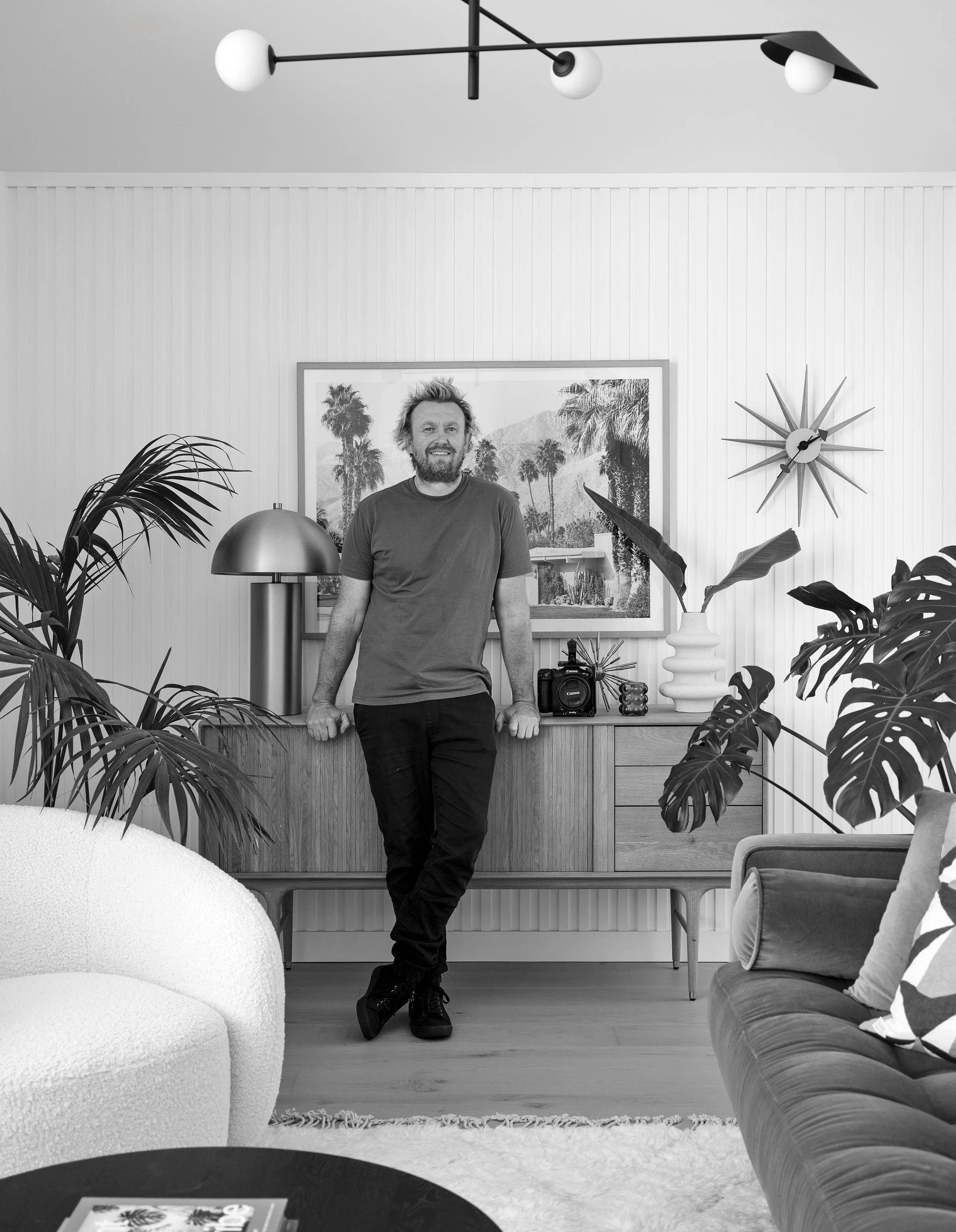 A man with a beard and casual clothing standing in a modern living room, leaning against a wooden sideboard, surrounded by plants, a lamp, and art.