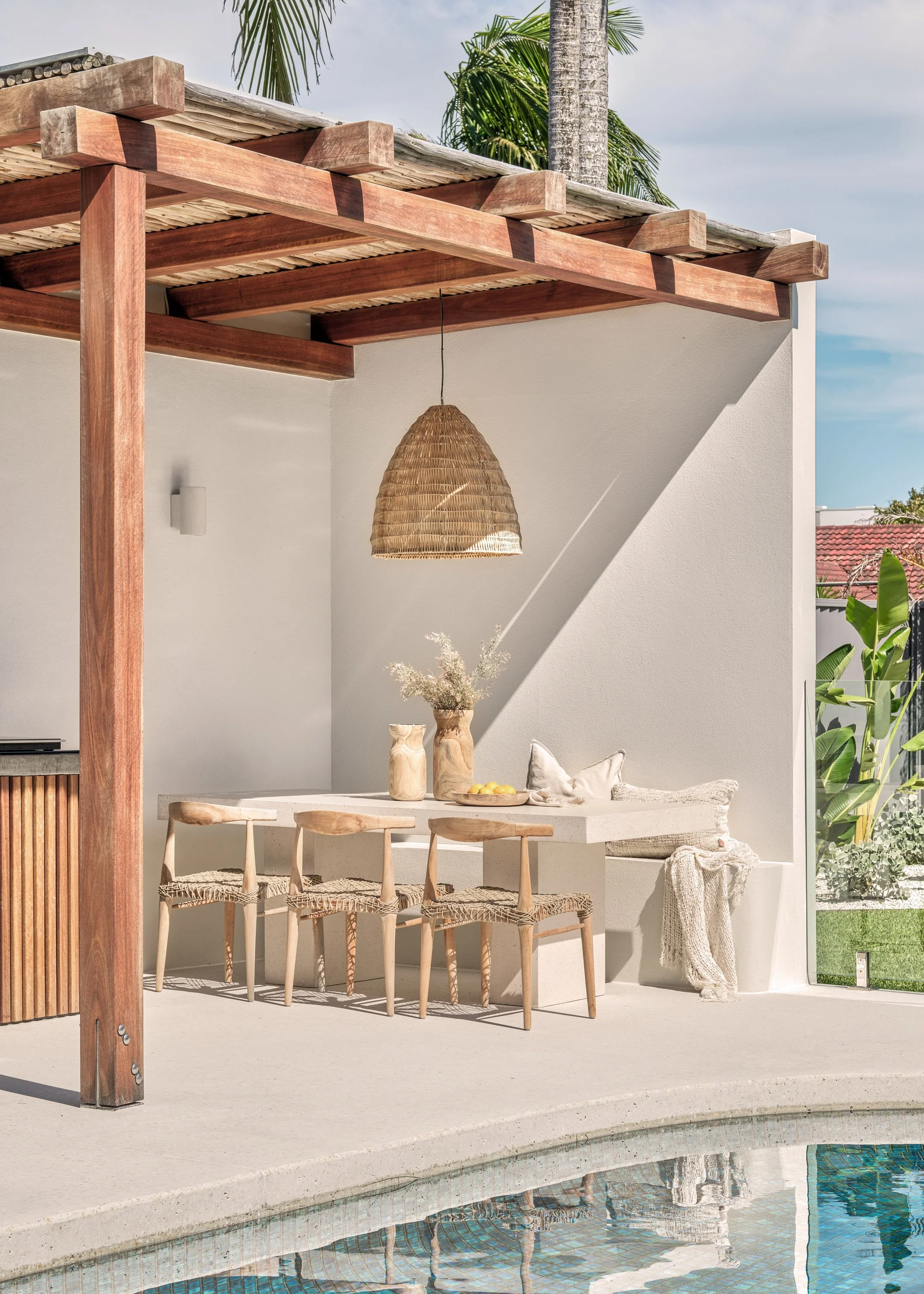 Outdoor patio area with a white table, three wooden chairs with woven seats, and decorative vases with dried flowers. A wicker pendant light hangs above, and a beige blanket and pillows are on a built-in bench along the white wall. There are palm tre