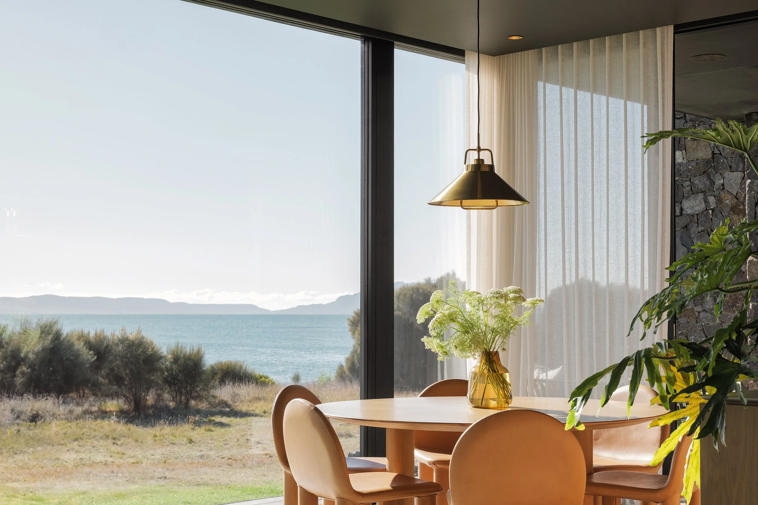 A dining area with a round wooden table, four caramel-colored chairs, a yellow vase with white flowers, and large glass windows revealing an outdoor landscape with trees, bushes, and water.