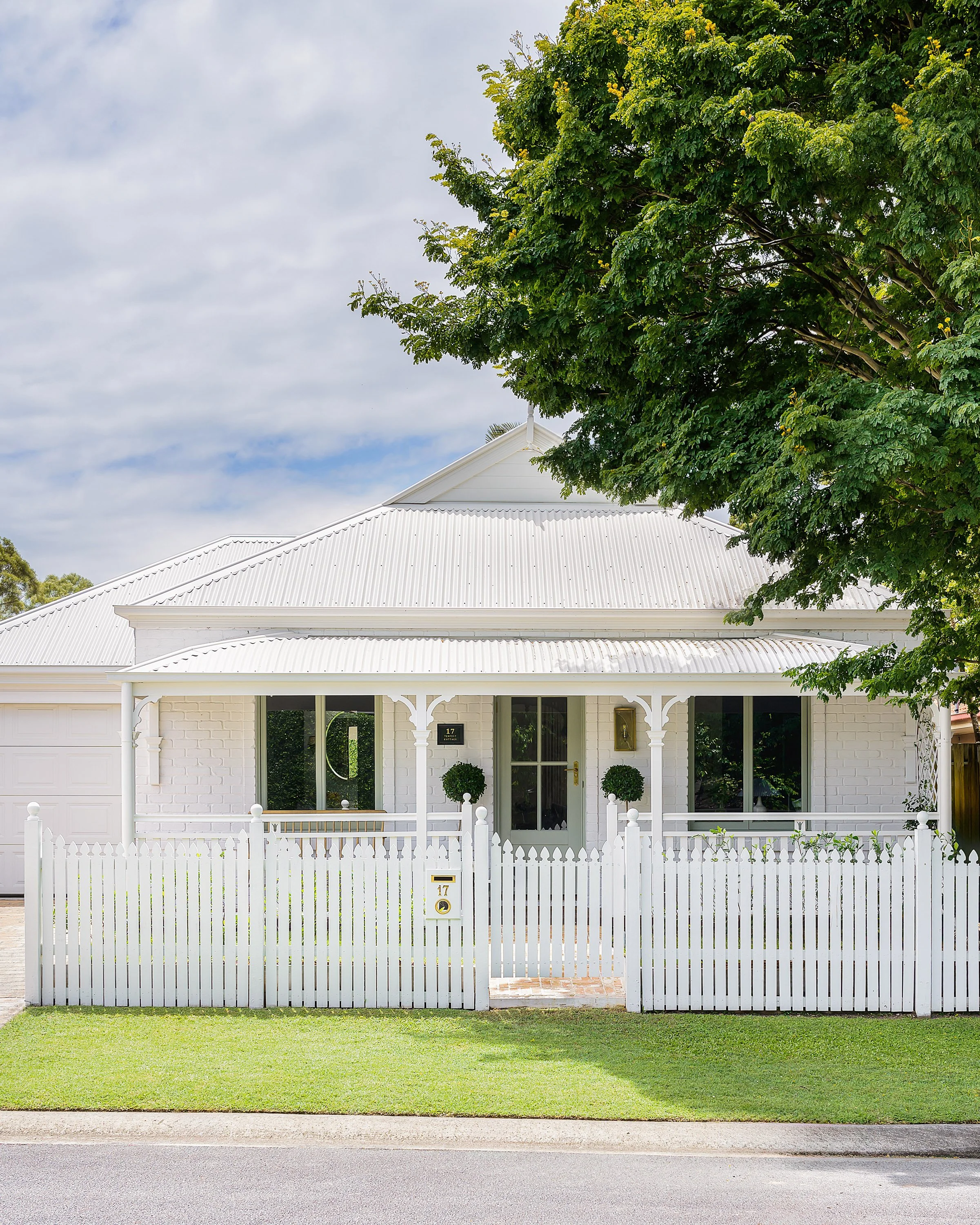 White house with a front porch, white picket fence, large tree, and manicured lawn.