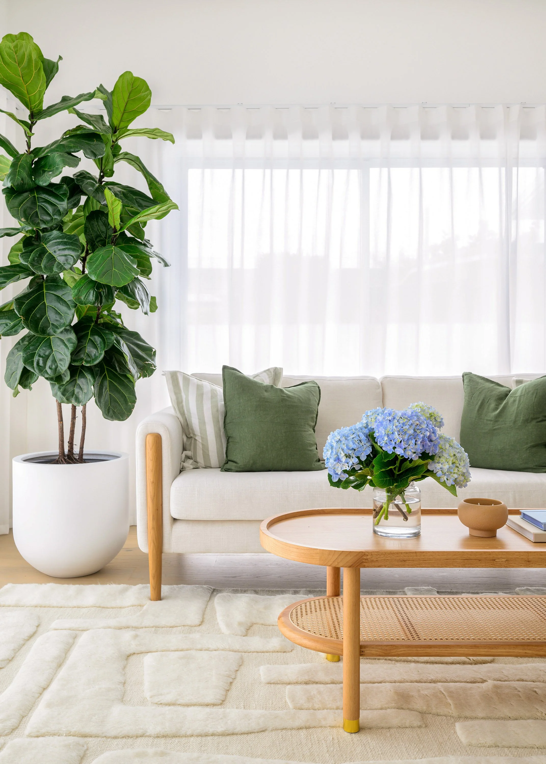 Living room with large l-shaped sofa, green and striped pillows, glass vase with blue hydrangeas, small wooden coffee table, large potted fiddle leaf fig plant, cream carpet, and white curtains.