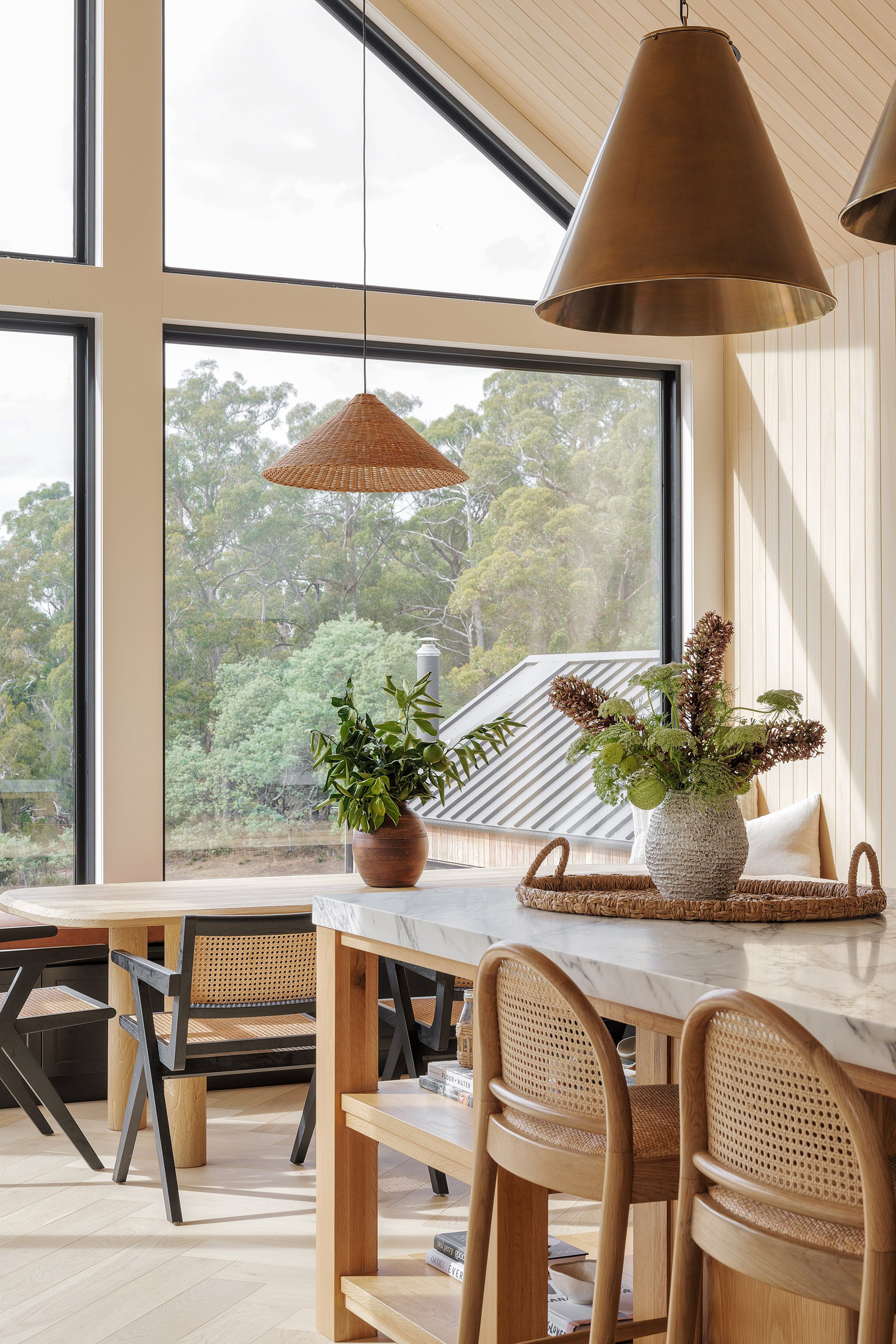 A cozy dining area with a marble-topped island, wooden chairs, potted plants, hanging pendant lights, and large windows showing trees outside.
