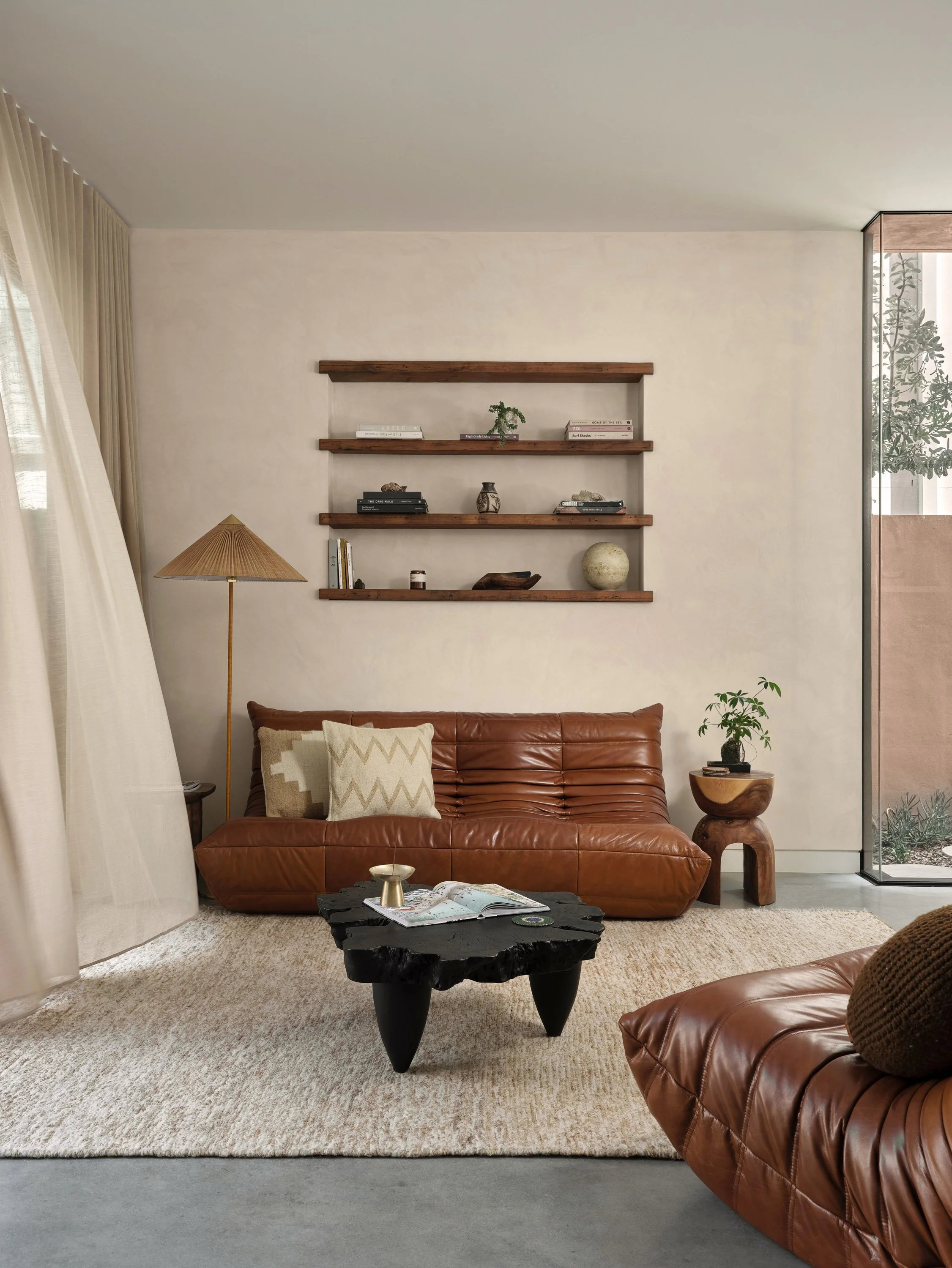 Modern living room with a brown leather sofa, a black coffee table, a side table with a plant, a tall floor lamp, a white textured wall with wooden shelves, and a large window with sheer curtains.