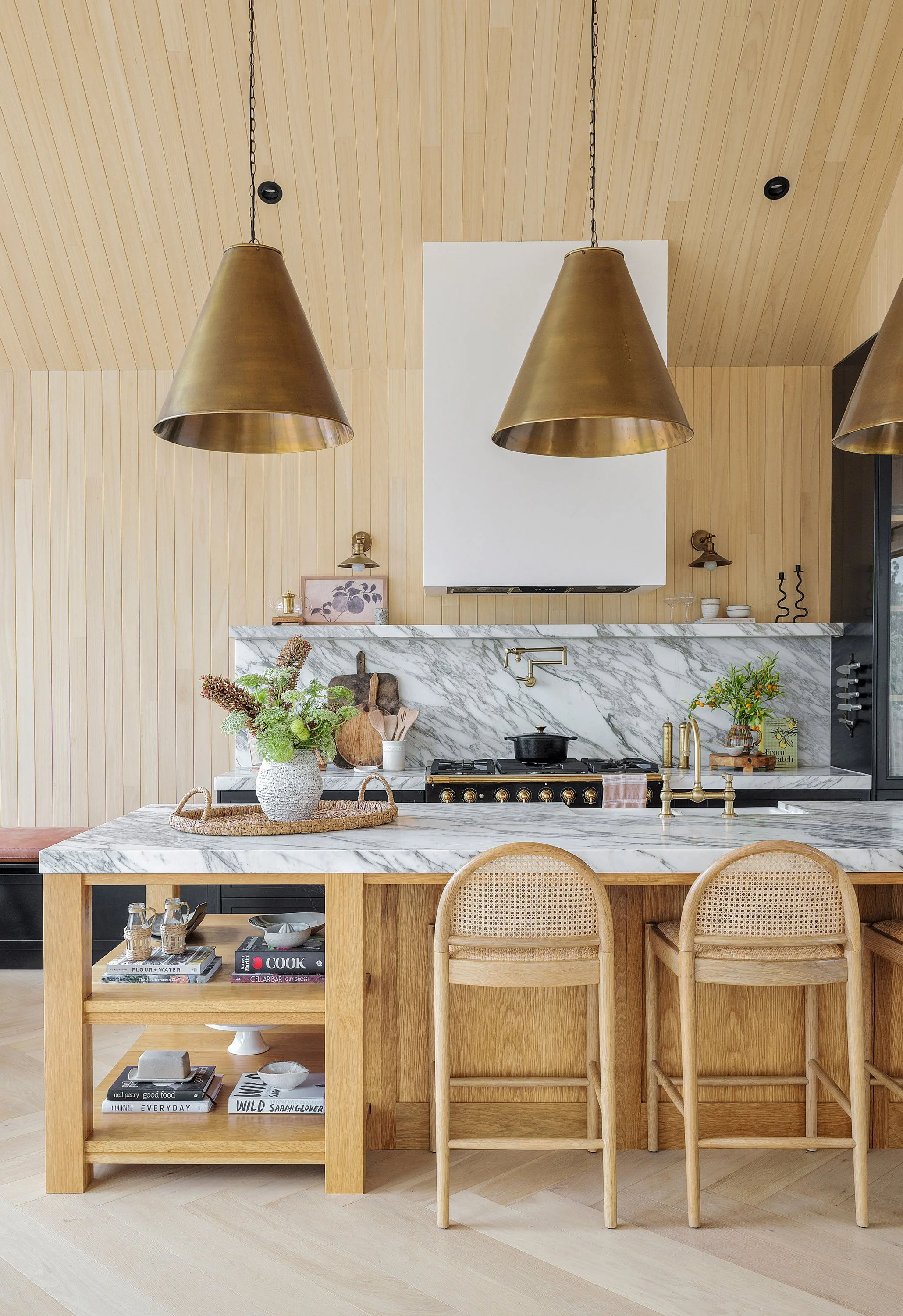 Modern kitchen with marble countertops, light wood cabinetry, and two large gold pendant lights hanging above a kitchen island. There are two rattan bar stools at the island and decorative items, plants, and books on the shelves and counters.
