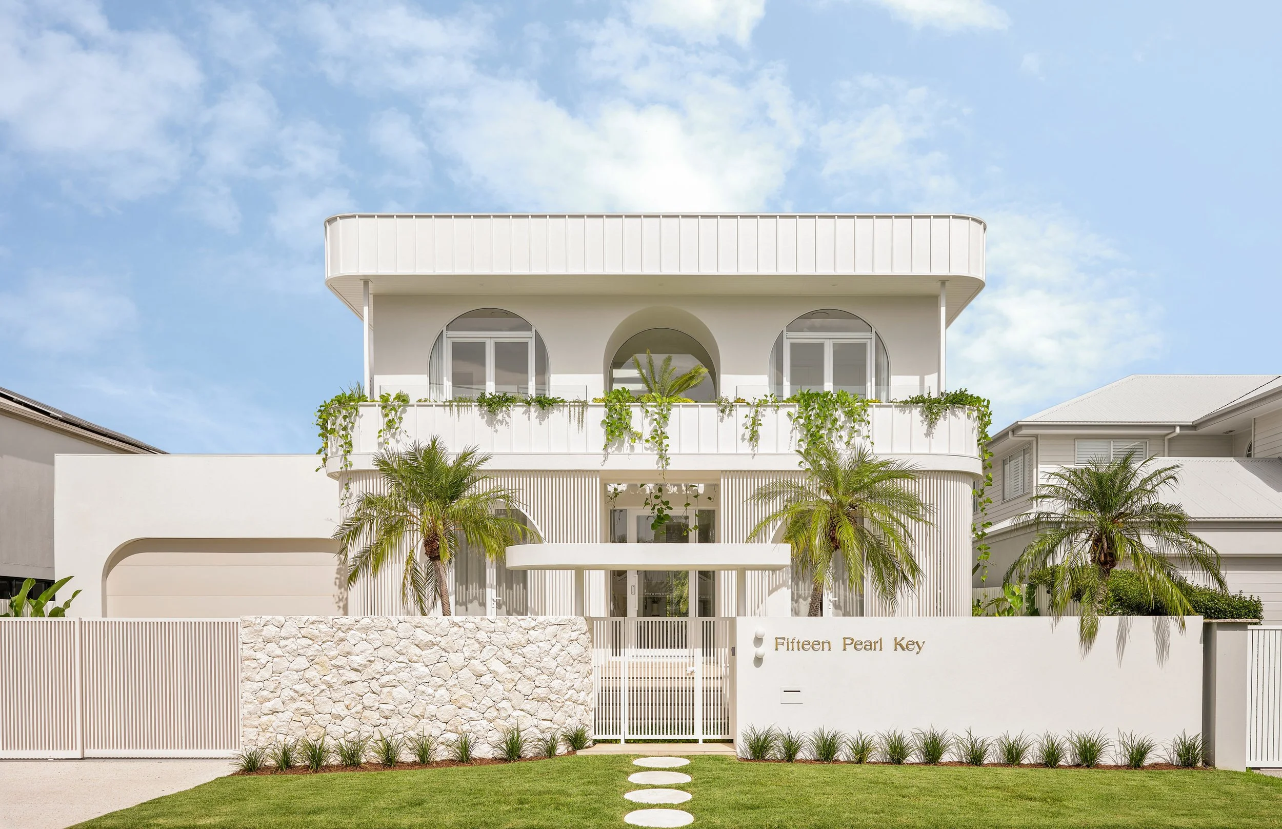 Modern white house with palm trees, a stone wall, gated entrance, and palm trees in front, under a blue sky.