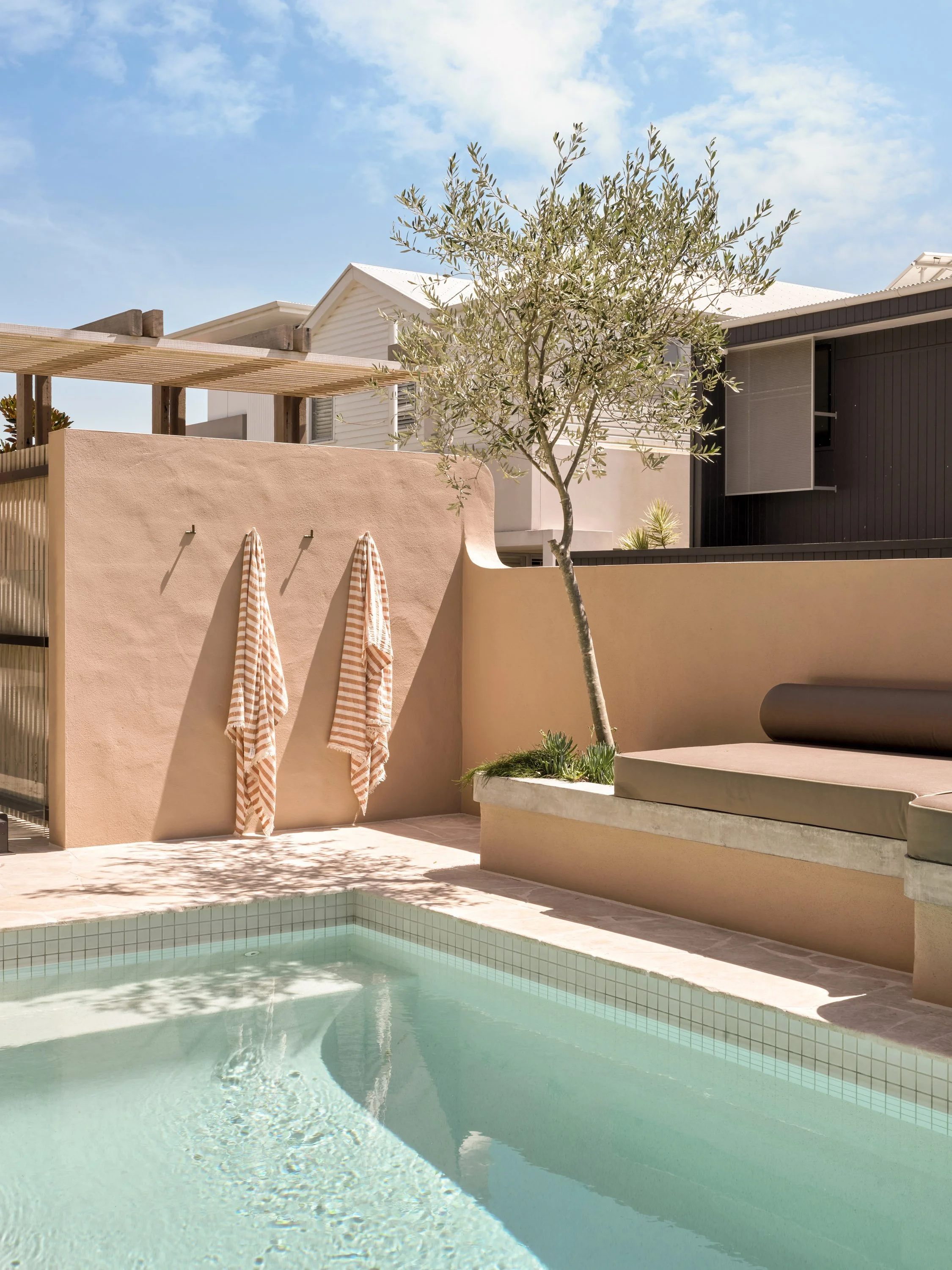 A modern backyard with a swimming pool, a small tree, two striped towels hanging on a beige wall, and outdoor seating with beige cushions under a clear blue sky.