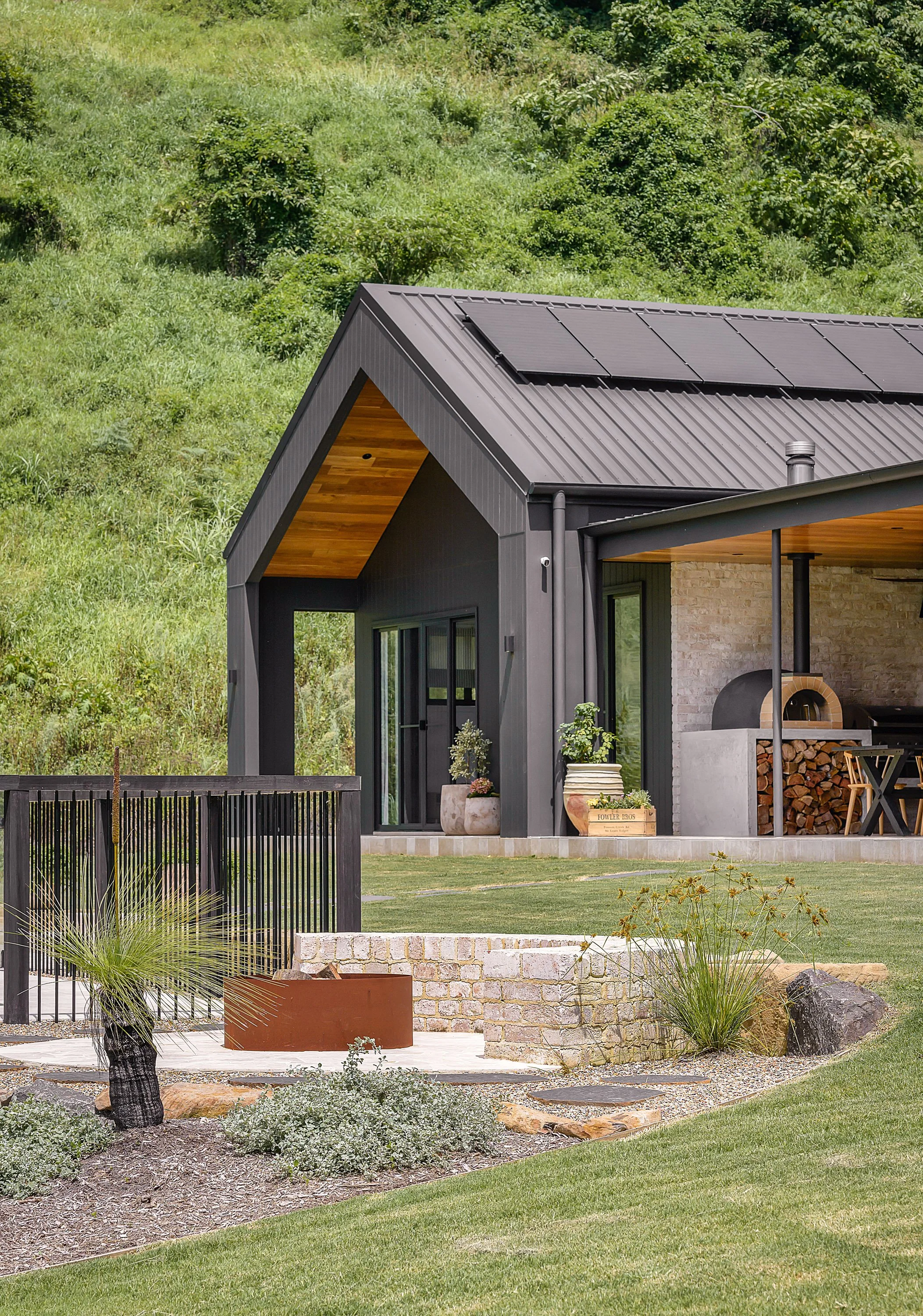 Modern house with dark exterior and wooden accents, surrounded by landscaped yard with plants, rocks, and grass, with a hillside in the background.