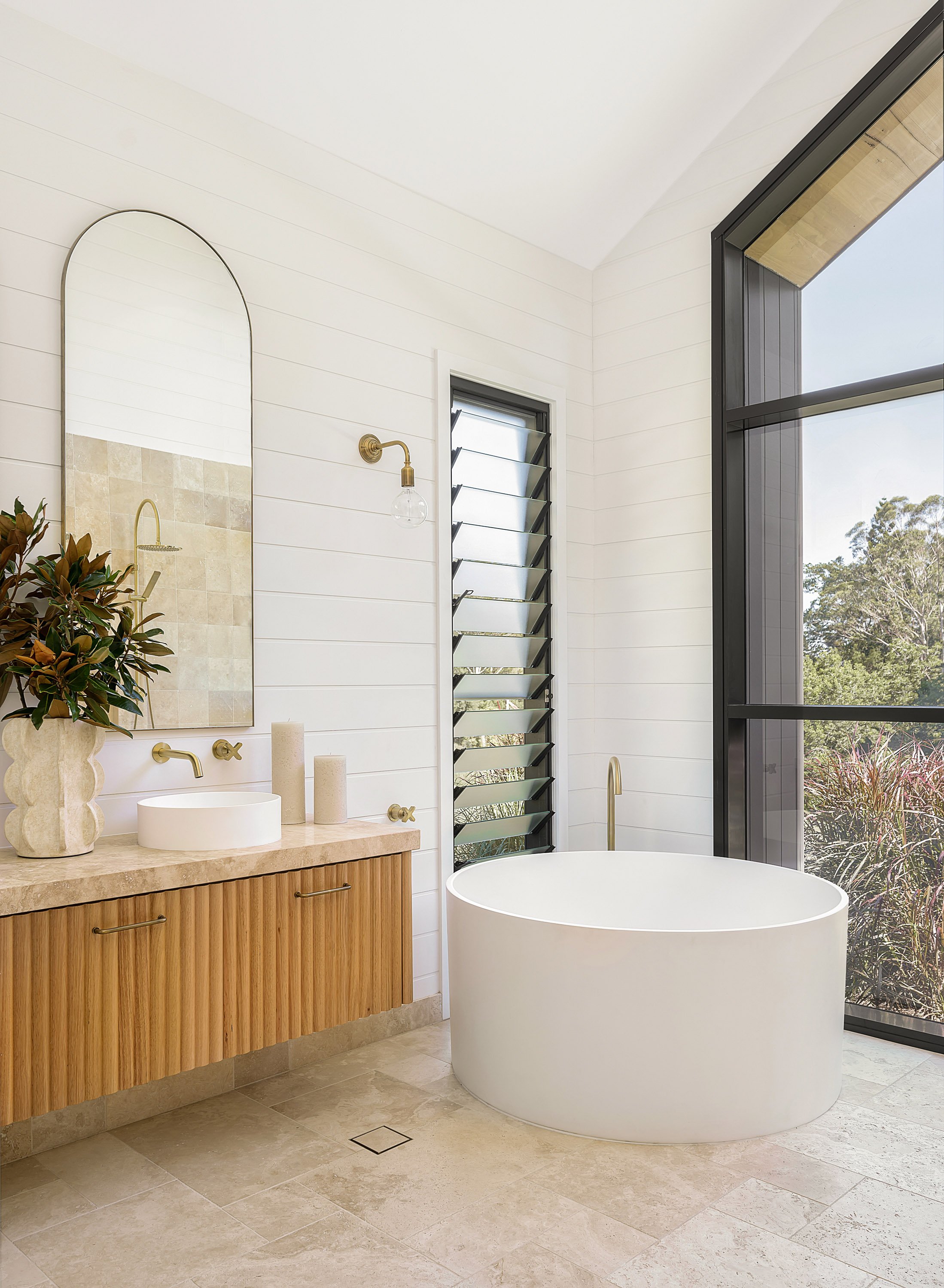 Modern bathroom with a round white bathtub, wooden vanity with a beige countertop, large window with black framing, and a mirror above the vanity. Decor includes potted plant and candles.