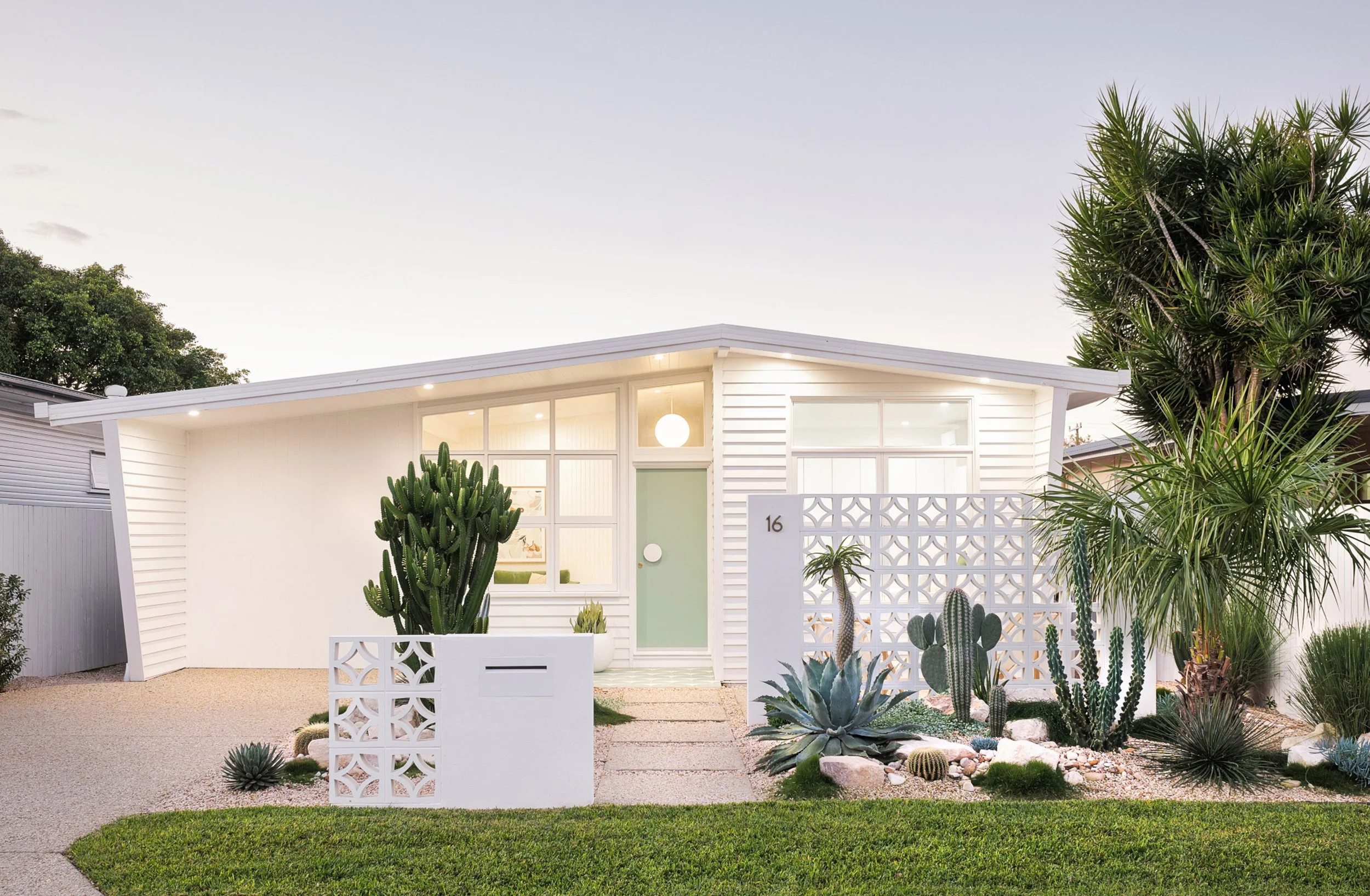 Modern white house with a garden featuring various cacti and succulents, a concrete pathway, and a white decorative fence, with soft lighting and a clear sky.