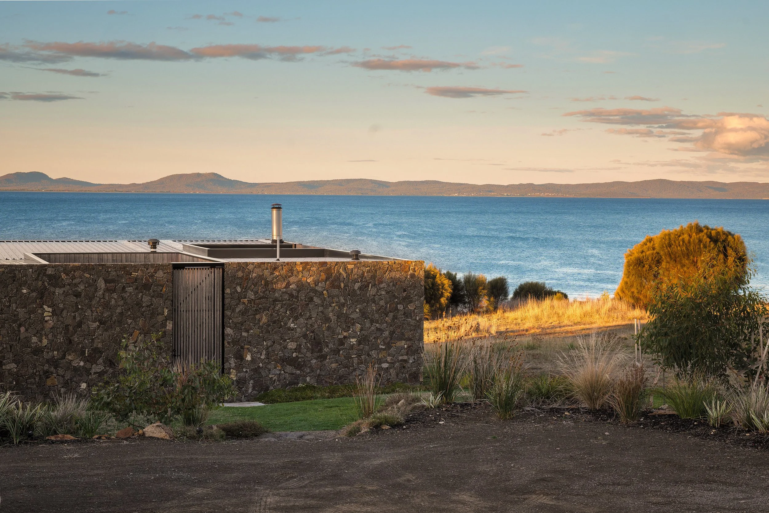 A modern stone building with a metal door, overlooking a large body of water with distant mountains, trees, and a cloudy sky at sunset.