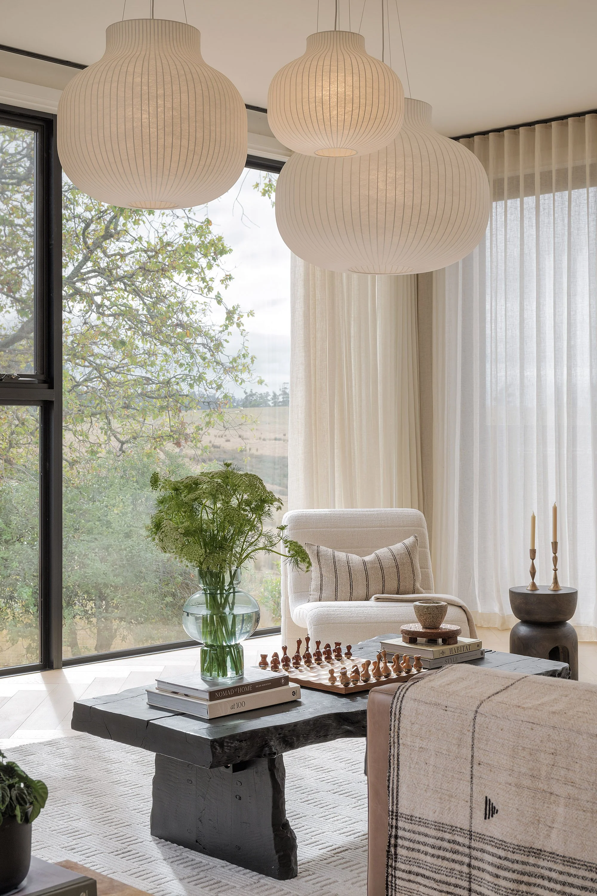 Living room with large window, white armchair, black wooden coffee table with books, a glass vase with green foliage, a chessboard, and decorative candles, with hanging paper lanterns and sheer curtains.