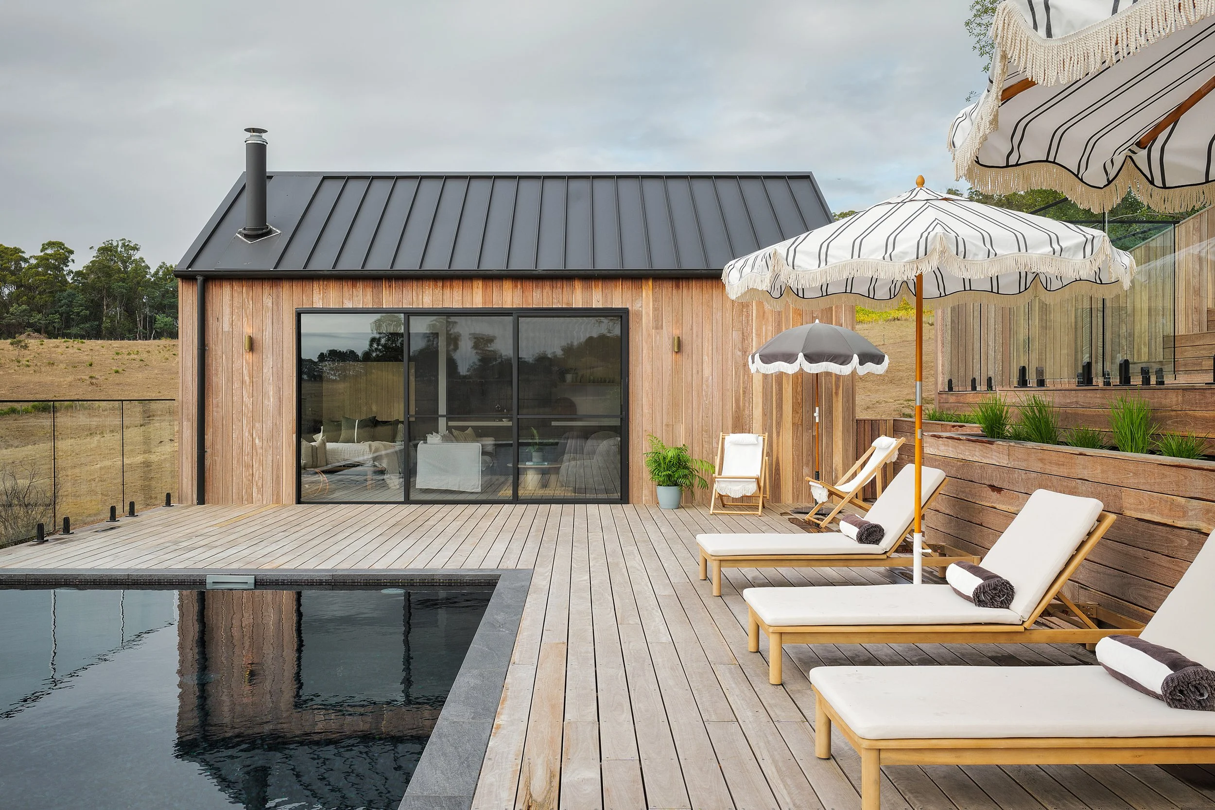 Outdoor patio with wooden deck, pool, white lounge chairs with black towels, large striped umbrellas, a small potted plant, and a modern wooden house with glass sliding doors and a metal roof, in a rural setting with open fields and trees in the back