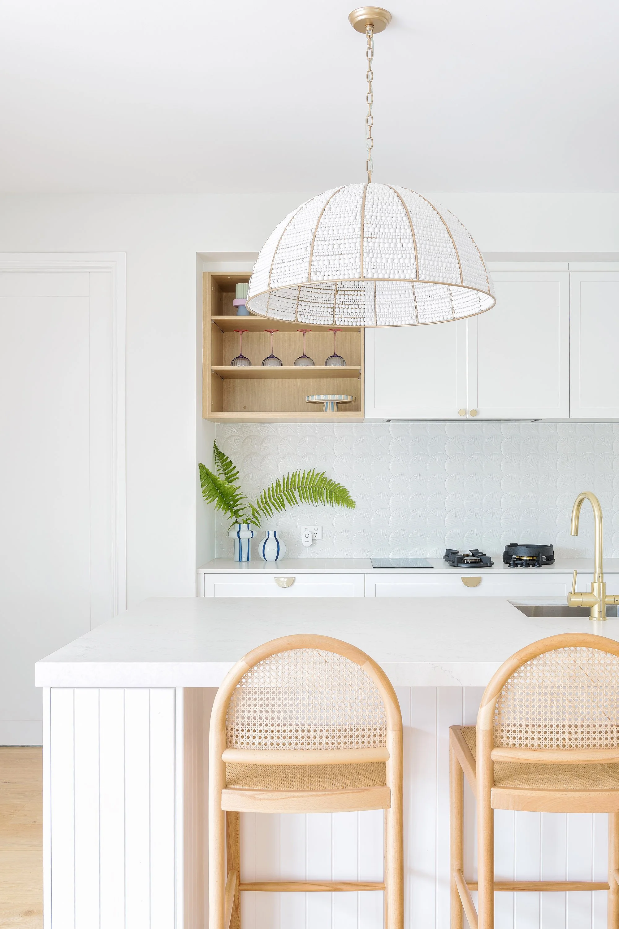 A modern, minimalist kitchen with white cabinets, a white countertop, and a gold faucet. Two light wood bar stools with cane backs sit at the counter. A pendant light with a white beaded shade hangs above. A small shelf with wine glasses and vases wi