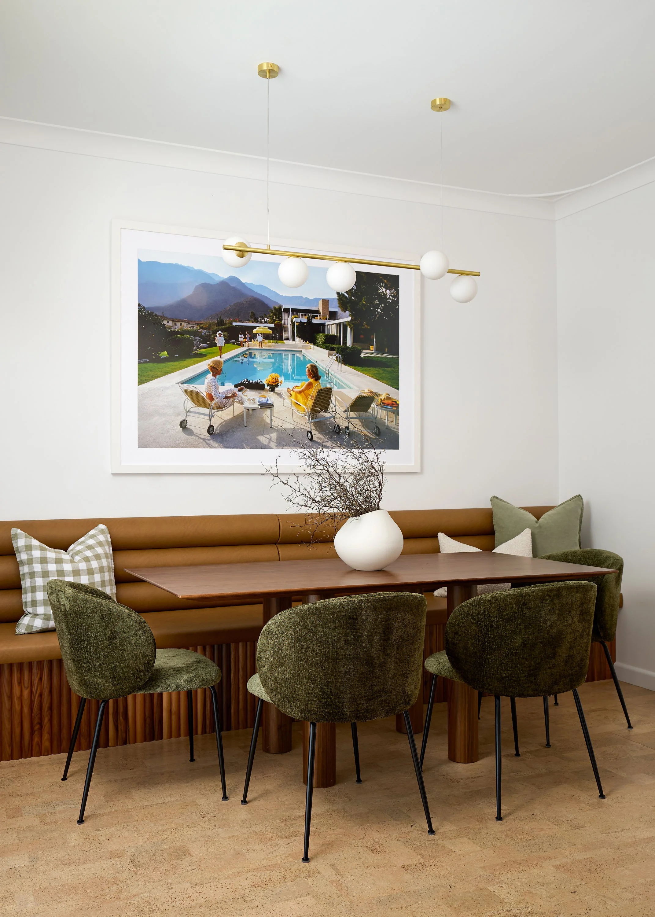 A dining room with a wooden table, green upholstered chairs, a tufted leather bench with checkered and plain cushions, and a white vase with twigs. A large framed photo of a poolside scene with people relaxing by the pool hangs on the wall. A modern 