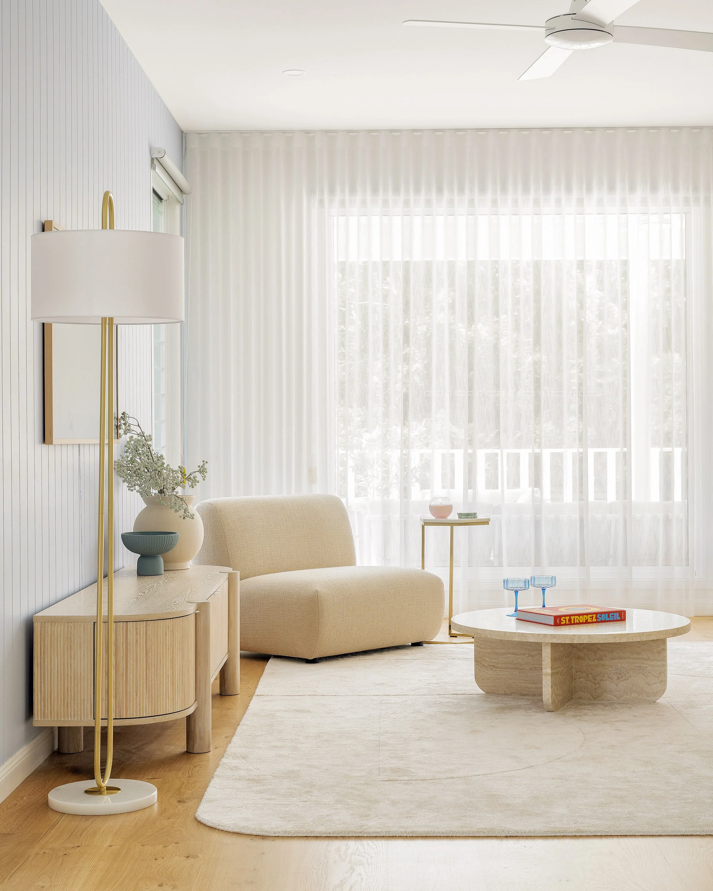 Bright living room with sheer white curtains, cream sofa, round marble coffee table with two blue glasses, a side table with a pink vase, and a tall floor lamp with a white shade and gold stand.