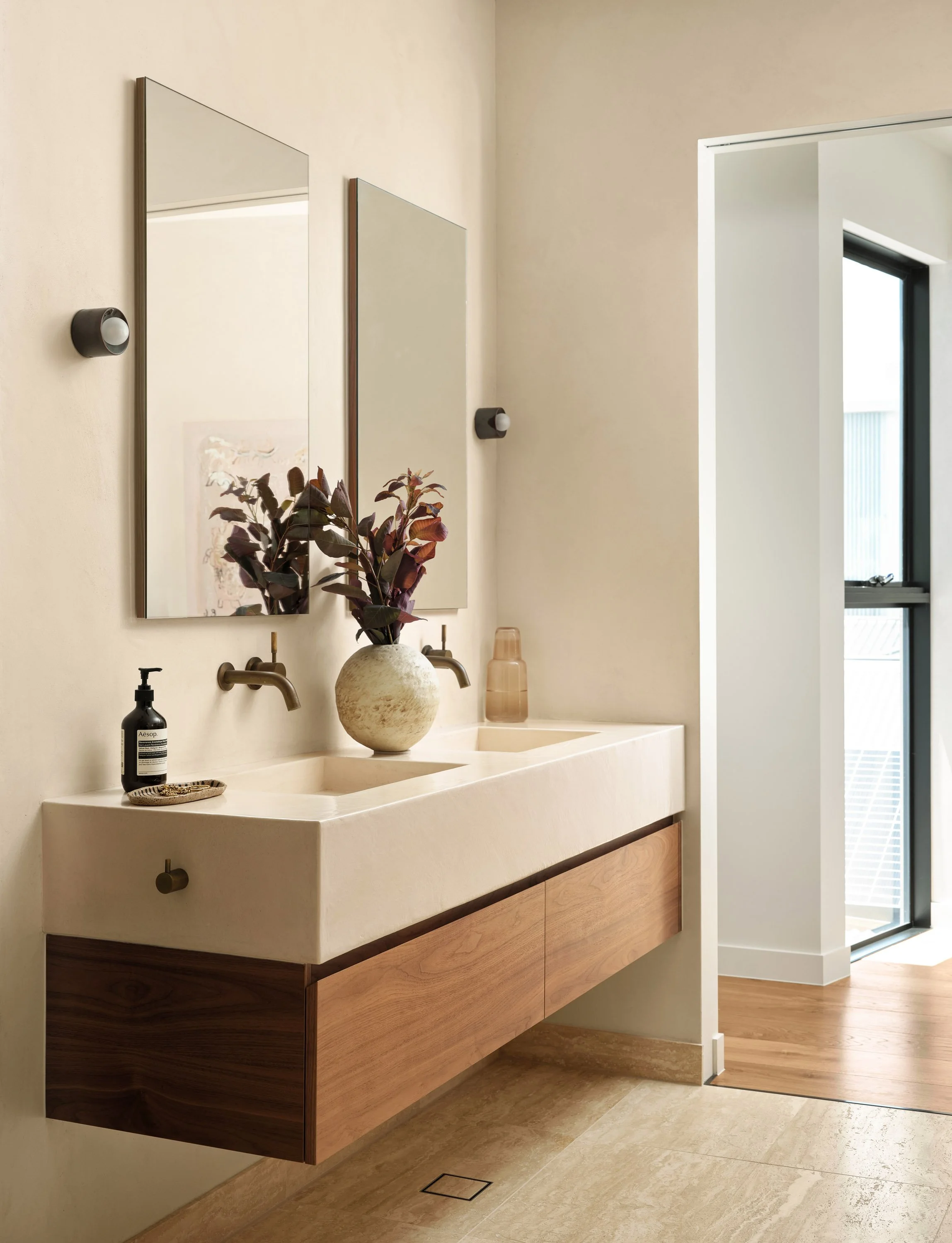 Modern bathroom with a floating vanity, double sinks, two mirrors, a decorative vase with dark leaves, and natural light from a window.