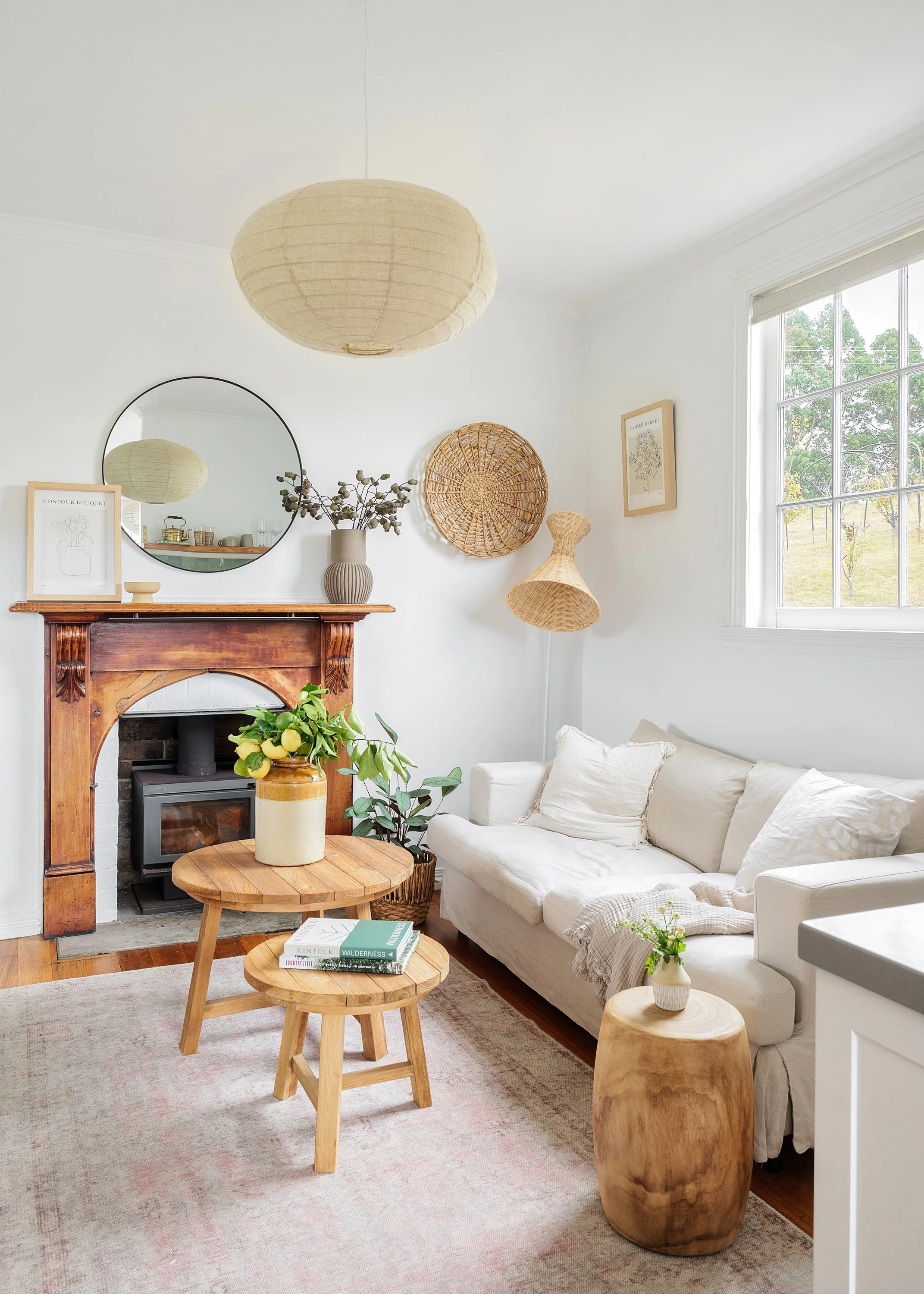 A cozy living room with a white sofa, wooden tables with books, a rustic fireplace mantel, a round mirror, and woven wall art and lighting decorations, illuminated by natural light from a large window.