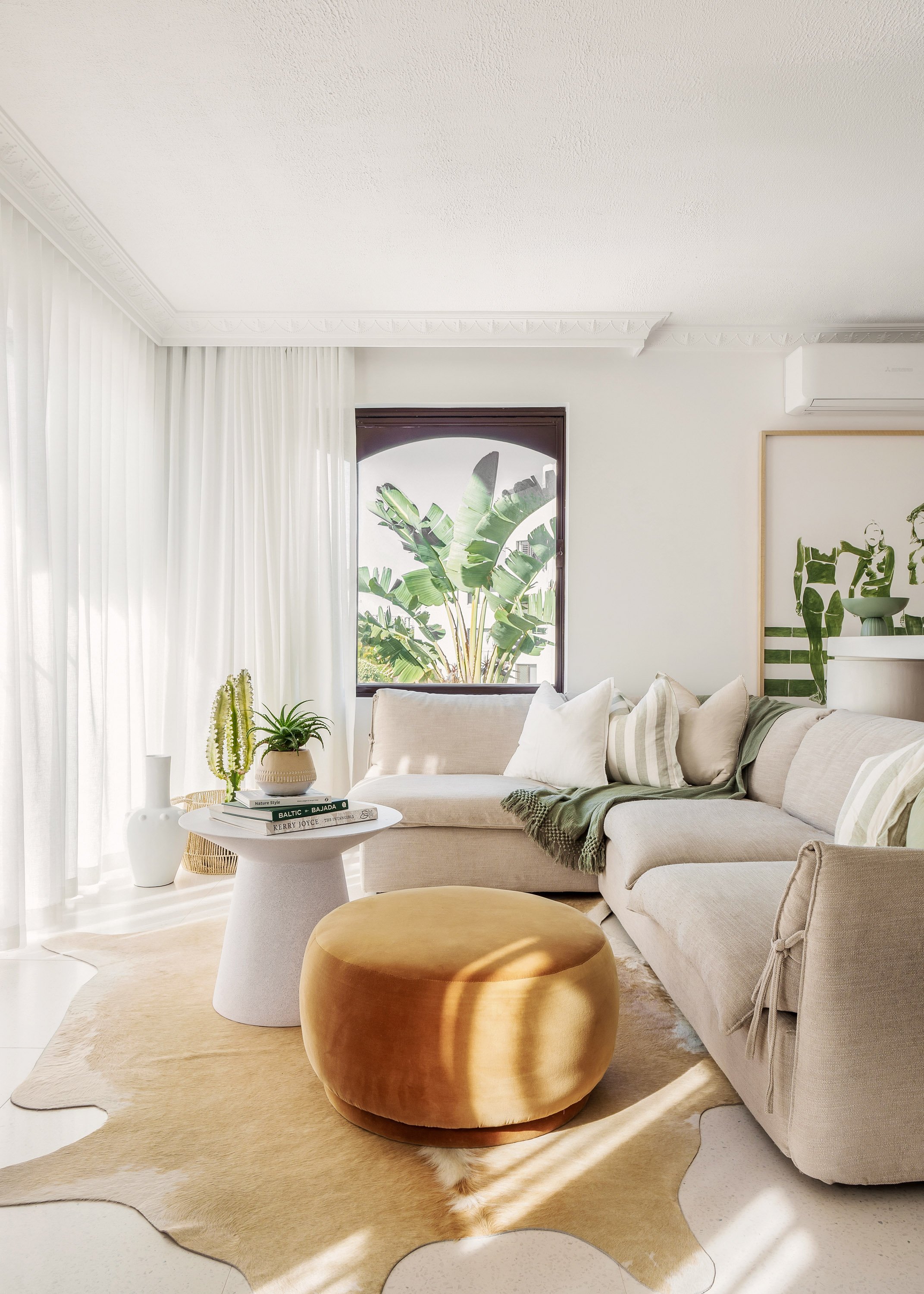 Bright living room with white curtains, a beige sectional sofa with pillows, a round ottoman, a white side table with houseplants and books, a large window with tropical plants outside, and modern artwork on the wall.