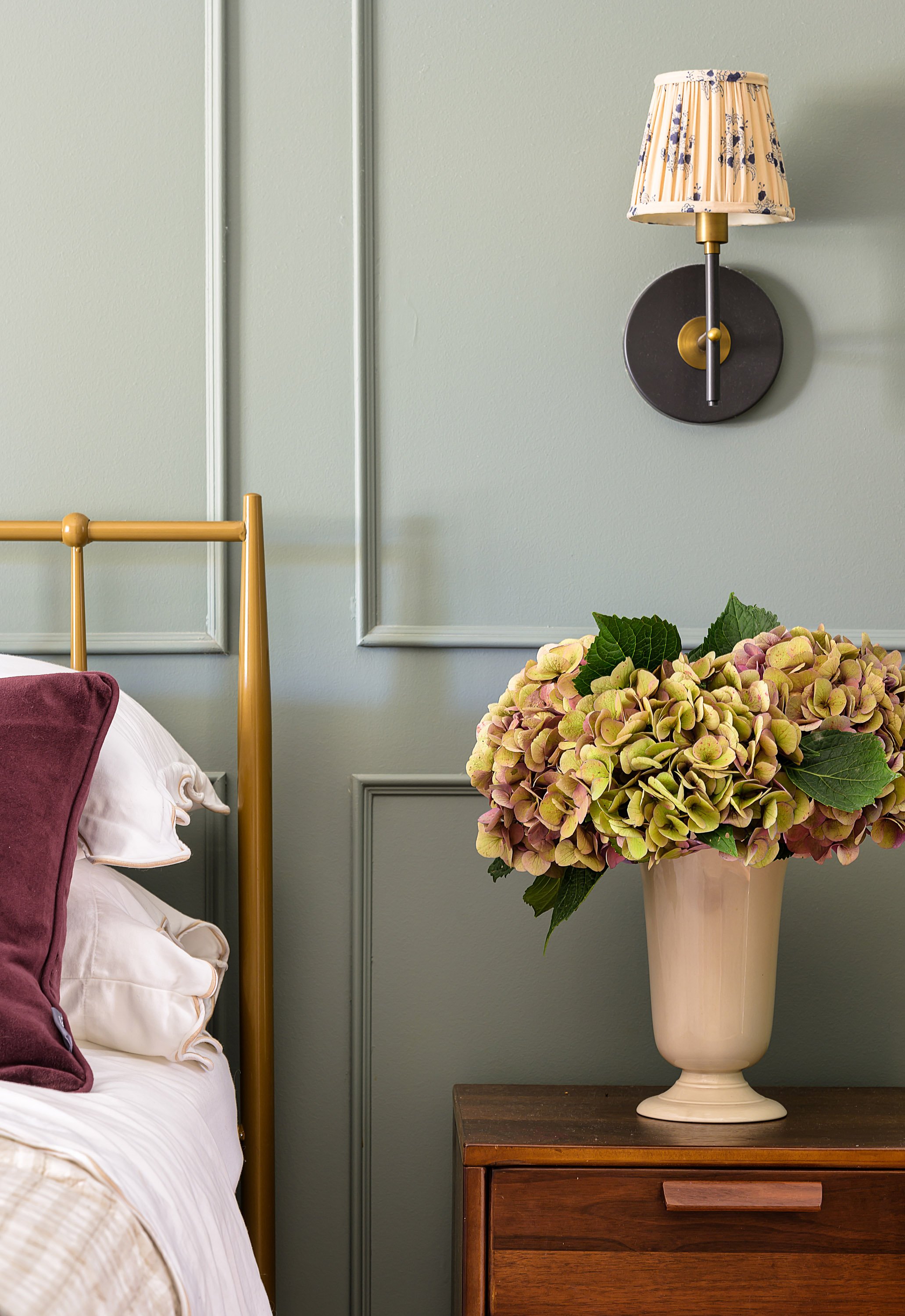 A bedroom corner with a gold bed frame, deep red accent pillow, white blanket, a wooden nightstand, a large flower arrangement with hydrangeas in a cream vase, and a wall-mounted lamp with a floral lampshade on a green wall.
