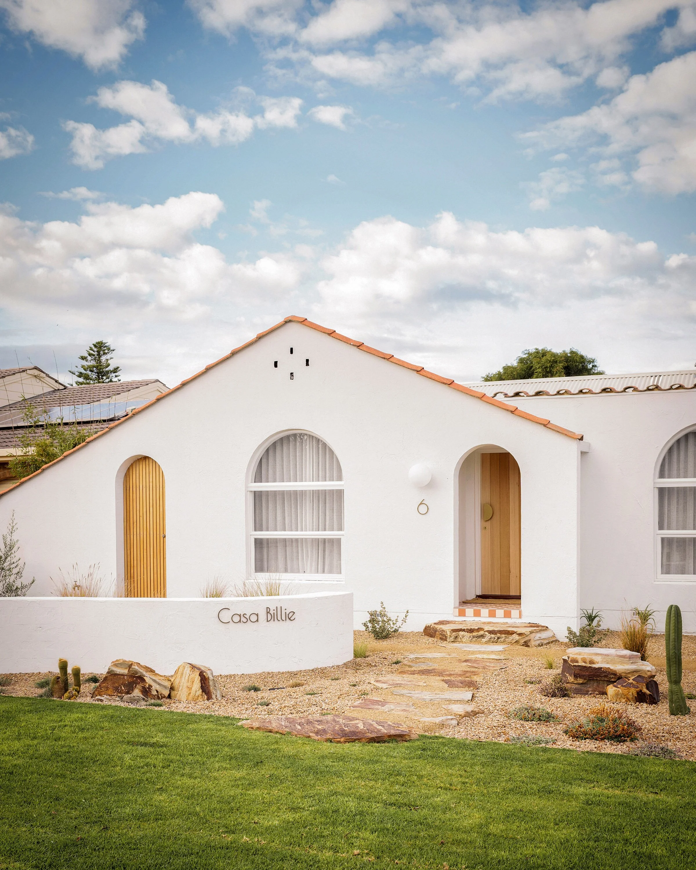 White modern house with arched windows and a wooden door, surrounded by desert plants and rocks, under a partly cloudy sky, with the words "Casa Billie" on a white wall in the front yard.