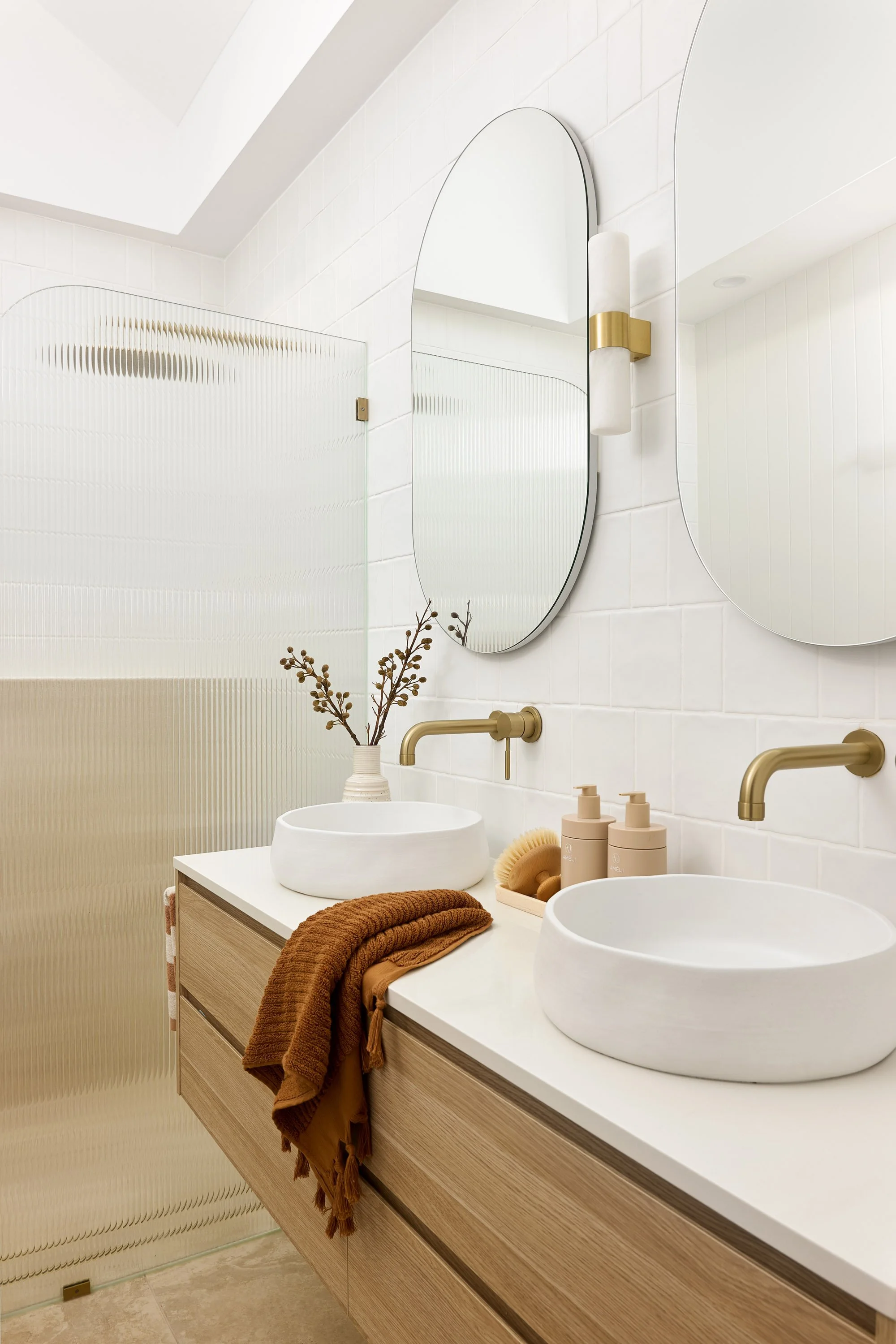 Modern bathroom with a double vanity, two white vessel sinks, gold fixtures, oval mirrors, a decorative vase with branches, soap dispensers, and a textured brown towel on the countertop.